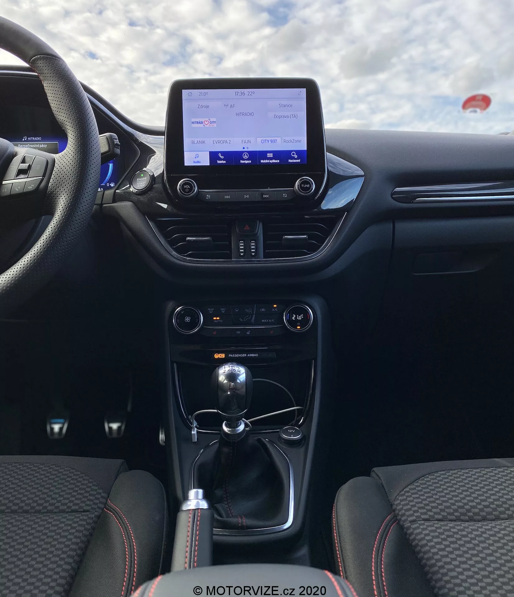 Vista interior del Ford Puma 2020 desde la perspectiva del pasajero trasero, enfocando en el área de la cabina central. La imagen resalta la consola central y el salpicadero, con una pantalla de infoentretenimiento, sistema de control de clima, palanca de cambios para transmisión manual y opciones de conectividad como puertos USB y toma de corriente de 12V. Los asientos están tapizados en tela con un patrón de costura distintivo y refuerzos laterales, enfatizando comodidad y deportividad.