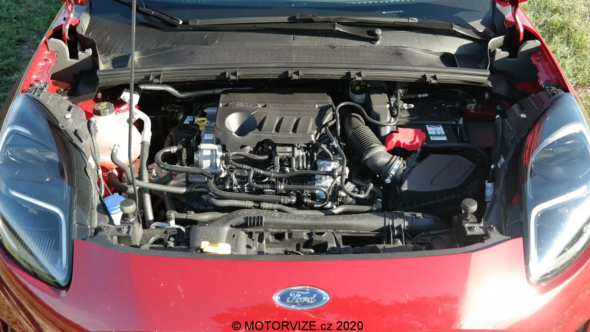 Vista detallada del compartimento del motor del Ford Puma 2020 con el capó abierto, mostrando componentes del motor como el bloque del motor, la tapa de válvulas, el colector de admisión y el colector de escape. También se pueden ver elementos visibles como depósitos de fluidos, mangueras, cables, batería, conductos de aire y parte del capó abierto con aislamiento.