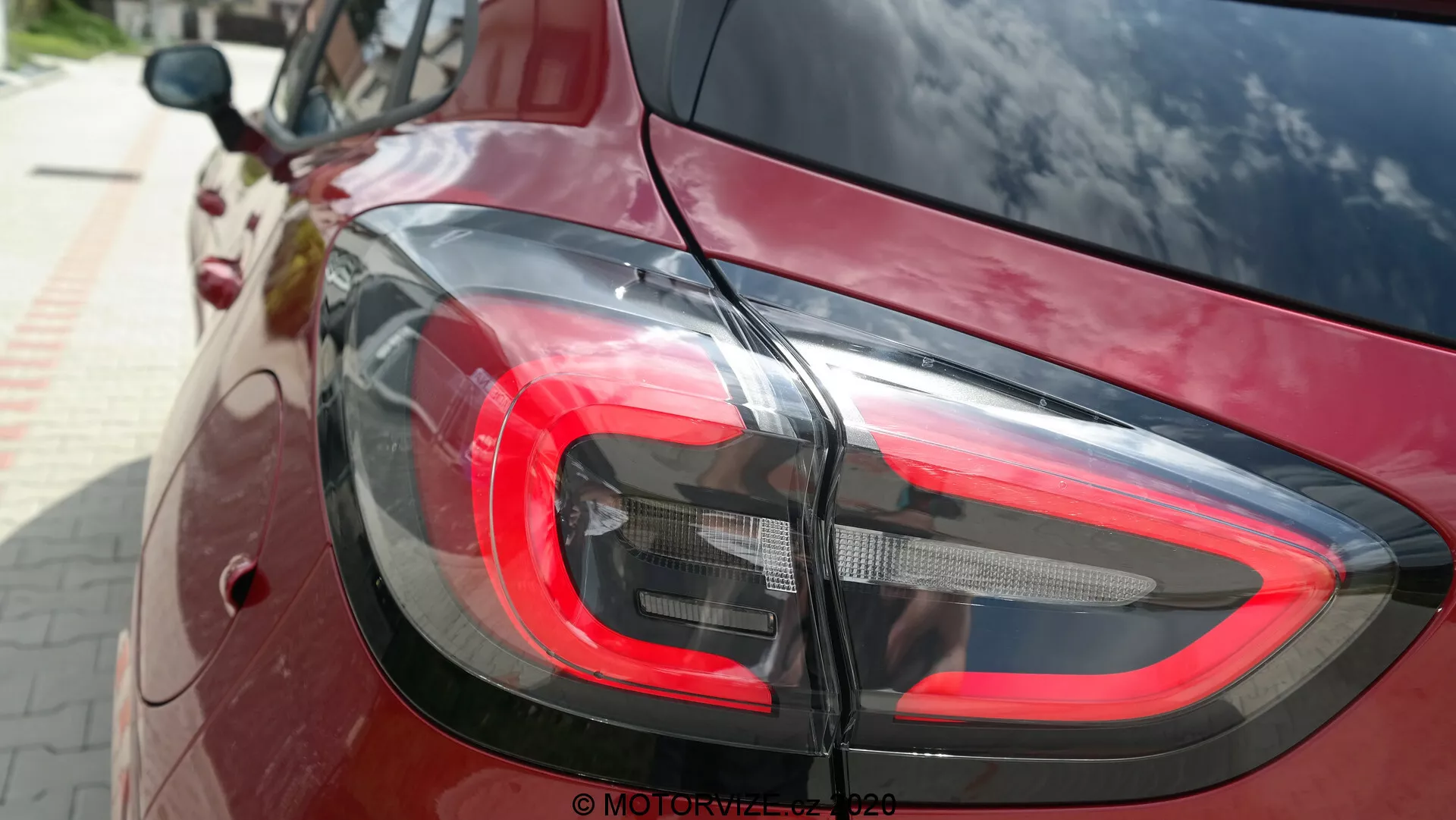 Primer plano de la sección trasera de un Ford Puma 2020, centrado en el diseño moderno de las luces traseras LED desde un ángulo ligeramente elevado. La luz de freno roja, los segmentos claros y oscuros para la luz de reversa y los indicadores de giro se mezclan perfectamente con la pintura de alto brillo de color rojo intenso o burdeos del automóvil, reflejando el entorno. La imagen resalta el estilo curvado y contemporáneo del automóvil y su tecnología de iluminación funcional.
