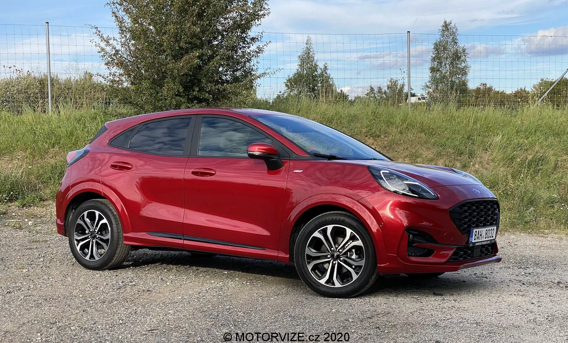 Fotografía profesional de un SUV crossover compacto Ford Puma 2020 rojo, posicionado en un terreno con hierba y follaje en el fondo. El ángulo lateral delantero destaca la prominente parrilla frontal con diseño de panal, faros LED elegantes y espejo lateral parcialmente visible. La línea de techo inclinada deportiva, las líneas distintivas de las puertas, manijas claramente visibles, ruedas de aleación con diseño de múltiples radios y neumáticos de bajo perfil contribuyen a la estética moderna del coche. La imagen, tomada a nivel del coche, captura toda su longitud, enfatizando su diseño contemporáneo.