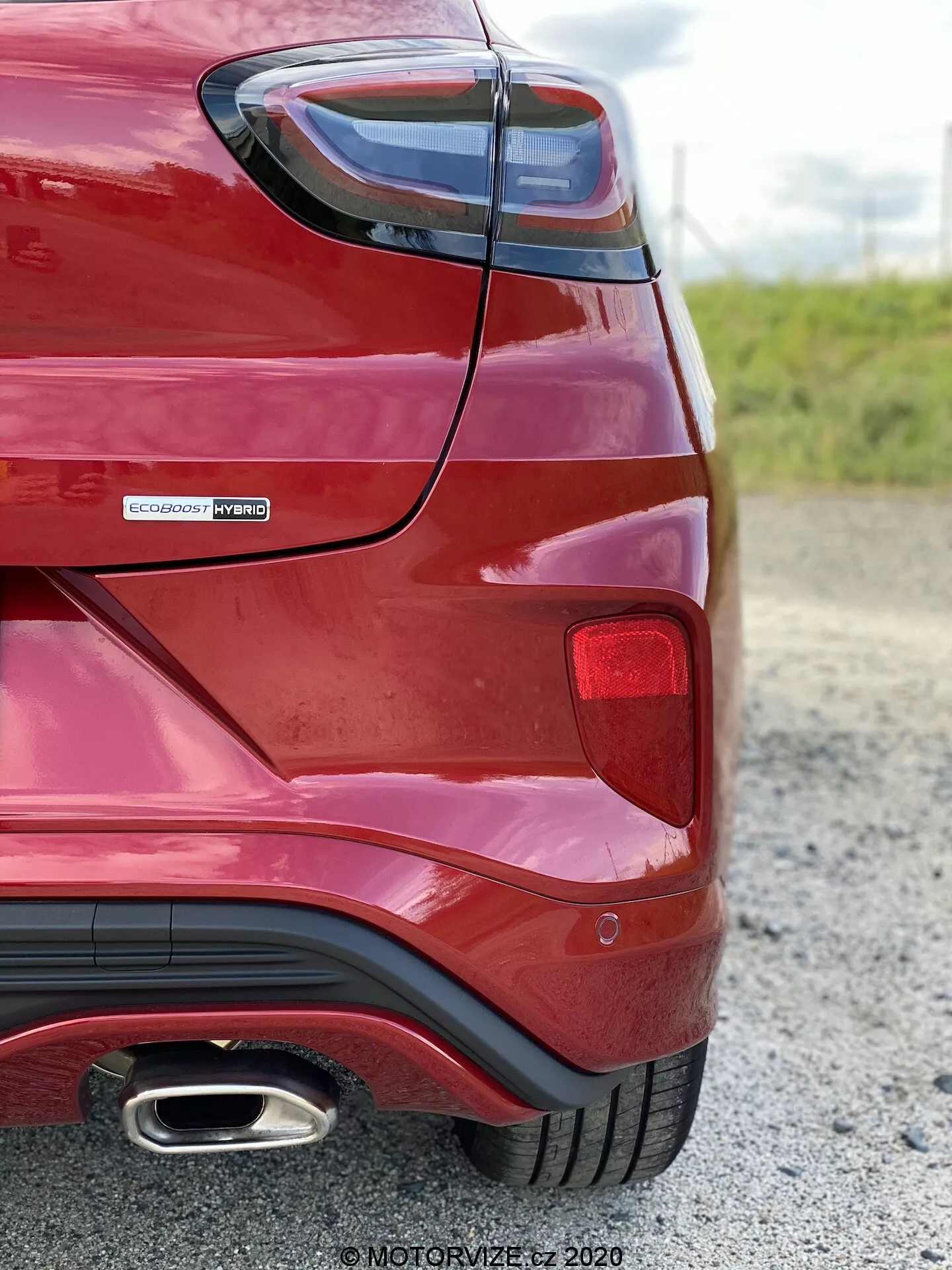 Vista de la esquina trasera de un Ford Puma 2020 rojo tomada desde un ángulo bajo y cercano, enfocándose en la curvatura del guardabarros trasero, el ensamblaje de las luces traseras, la insignia EcoBoost Hybrid que indica un tren motriz híbrido y la única punta de escape cromada. Resaltando elementos de diseño modernos como luces traseras en ángulos agudos y parachoques trasero aerodinámico con estilo de difusor.