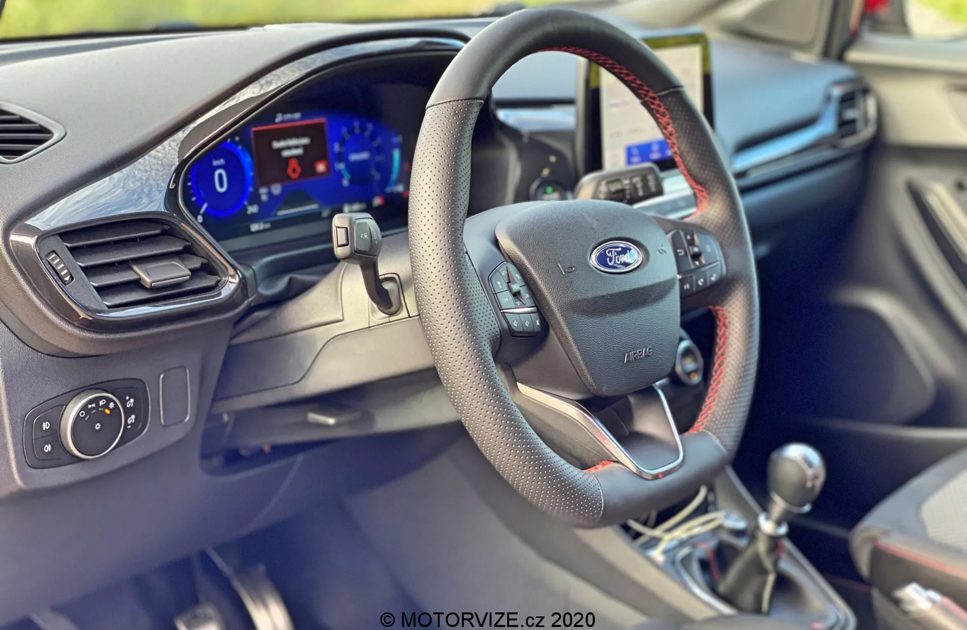 Vista interior del Ford Puma 2020 desde la perspectiva del asiento del pasajero a nivel de los ojos, destacando el lado del conductor. Características prominentes incluyen un volante negro con costuras rojas, con el emblema de Ford y botones de control modernos. El panel de instrumentos digital con una iluminación azul brillante es visible en el tablero. La perilla de cambios en la parte inferior derecha, con costuras, se encuentra sobre una palanca de cambios de transmisión manual. Parte de la puerta del conductor muestra controles montados en la puerta, una ventilación de aire y configuraciones adicionales, indicando un diseño automotriz y tecnología sofisticados.