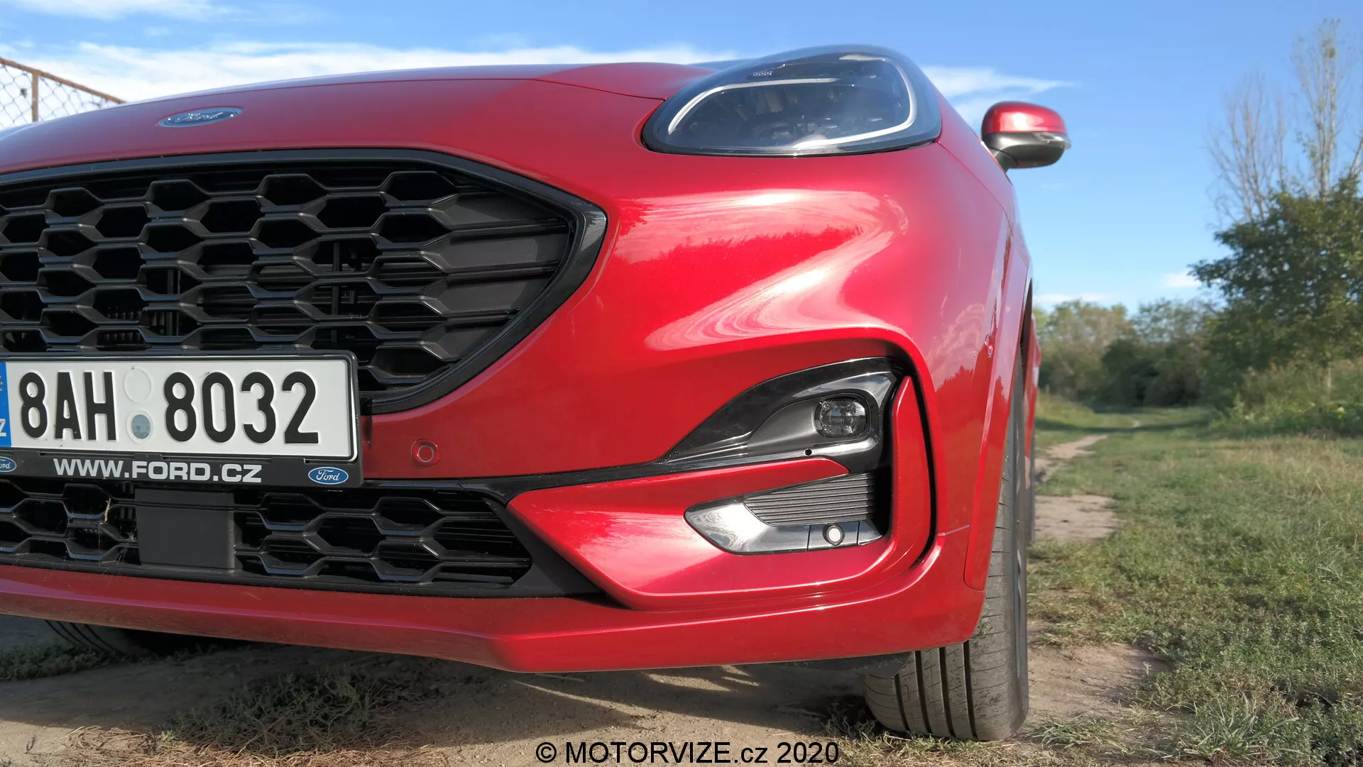 Vista en ángulo y primer plano de un Ford Puma 2020 rojo enfocado en el cuarto frontal izquierdo. Las características incluyen rejilla en forma de panal, faro LED/HID elegante, parachoques delantero y capó aerodinámico esculpido. Se ve una matrícula europea con una dirección web checa. Se observan parcialmente elementos modernos como las señales de giro integradas en el espejo lateral. La imagen, tomada desde un ángulo bajo, usa luz natural y poca profundidad de campo para resaltar el diseño del coche en un entorno al aire libre.