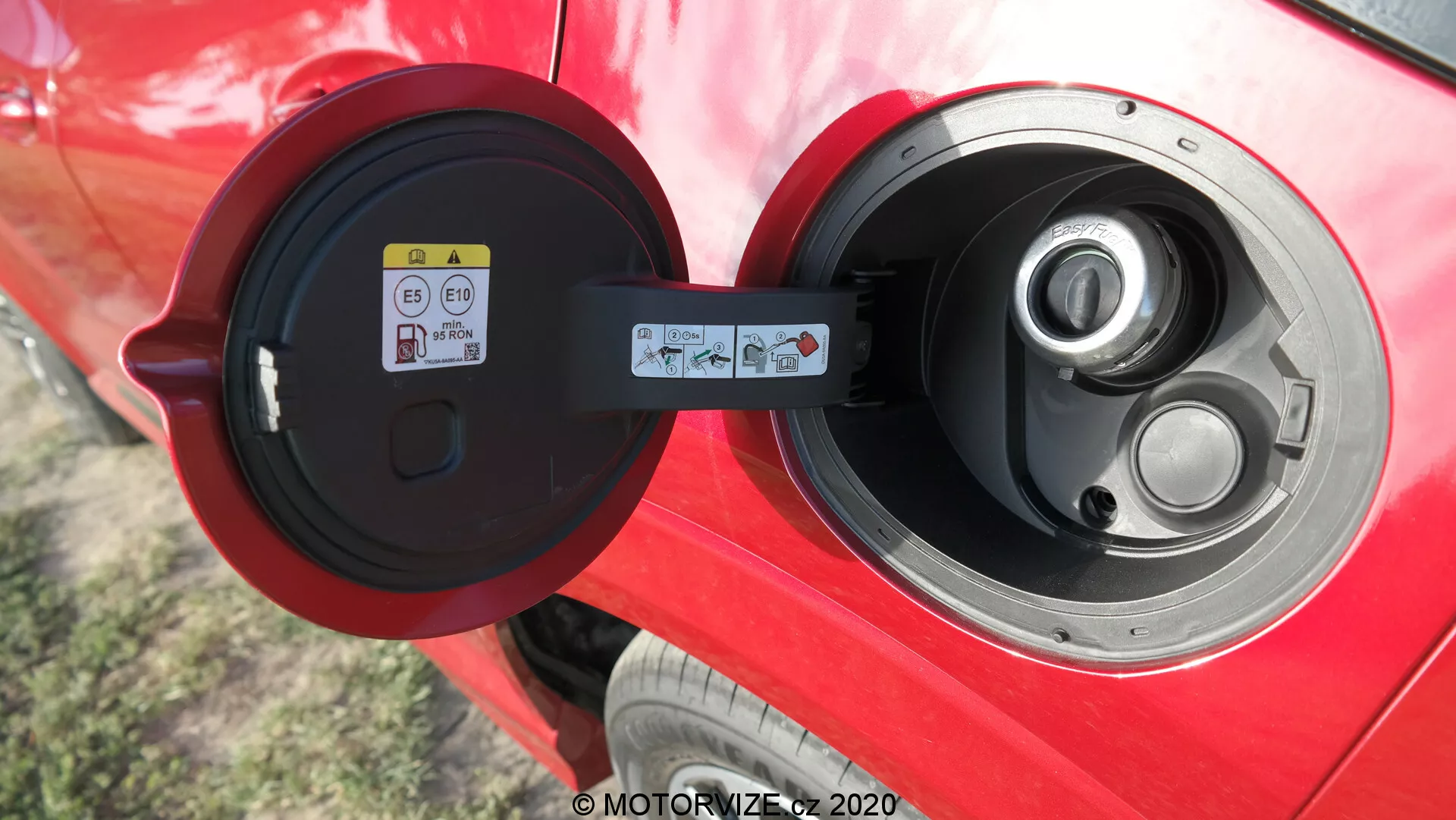 Vista de cerca de un puerto de carga tipo 2 abierto en un vehículo eléctrico o híbrido enchufable de color rojo vibrante, con un enfoque en el área del puerto brillante, la tapa con bisagras y una pegatina informativa visible a la izquierda dentro de la puerta del puerto, lo que sugiere un automóvil bien mantenido con tecnología avanzada de vehículos eléctricos en el exterior.