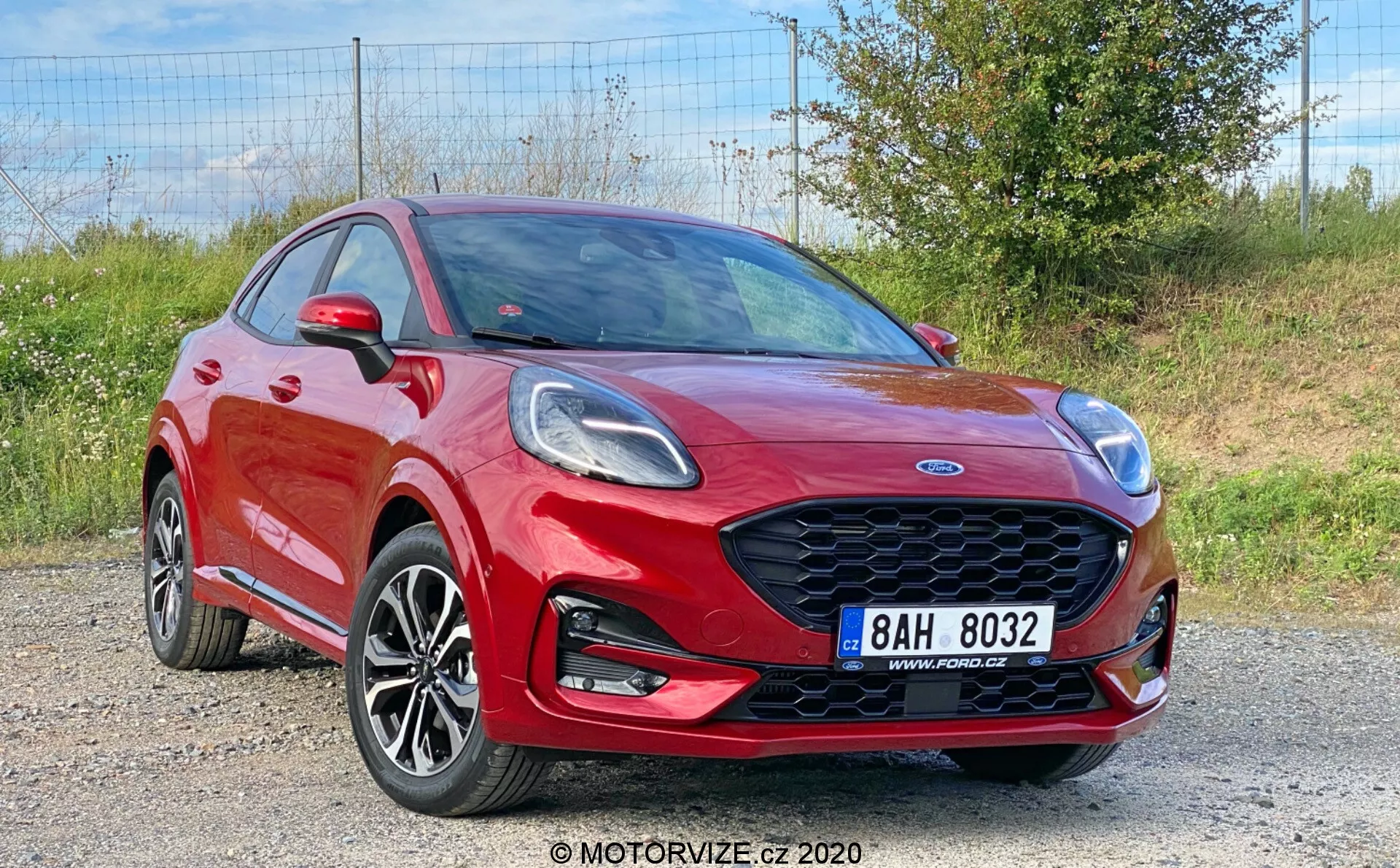 La imagen muestra un Ford Puma 2020 rojo vibrante, un vehículo crossover capturado desde una vista tres cuartos frontal, mostrando su elegante parrilla delantera de panal, faros angulares, llantas de aleación de múltiples radios, parachoques delantero con luces antiniebla integradas y una línea de hombro pronunciada. El capó dinámico con pliegues, espejos laterales con indicadores de giro y parte de las puertas delanteras y traseras son visibles, lo que indica una configuración de cuatro o cinco puertas, en un fondo de grava con vegetación.