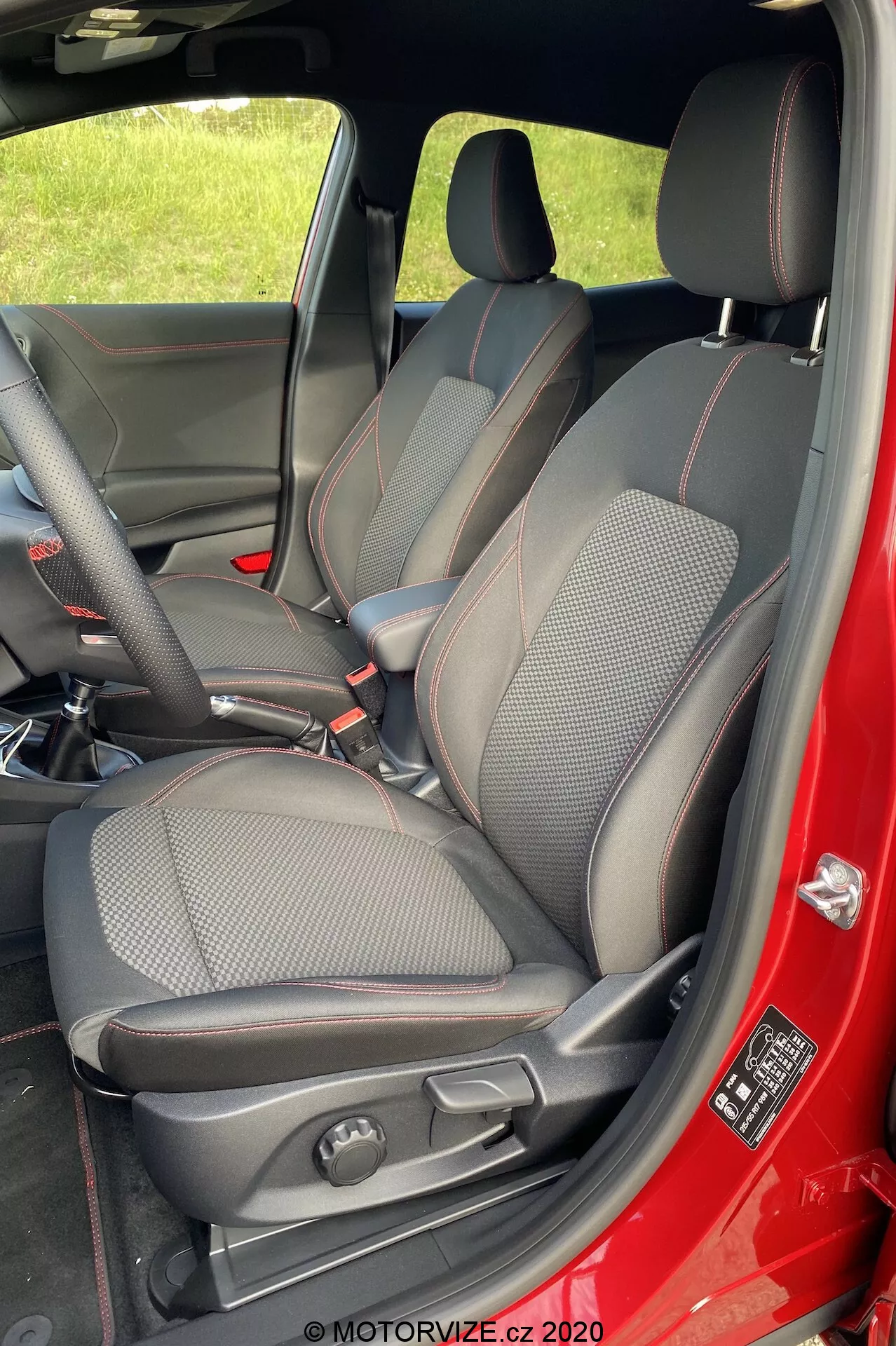 Imagen vertical del interior del Ford Puma 2020 enfocada en el lado del pasajero delantero. Tomada desde afuera con la puerta abierta, mostrando completamente el asiento del pasajero delantero, partes del asiento del conductor, la consola central y el interior de la puerta. El asiento tiene un diseño deportivo con una mezcla de cuero y tela, costuras contrastantes rojas, reposacabezas integrado y soportes laterales. Los controles de ajuste manual del asiento son visibles. El color rojo del exterior del auto se refleja en el borde de la puerta y el panel interior de la puerta es negro. En el marco de la puerta hay una etiqueta de seguridad e información.