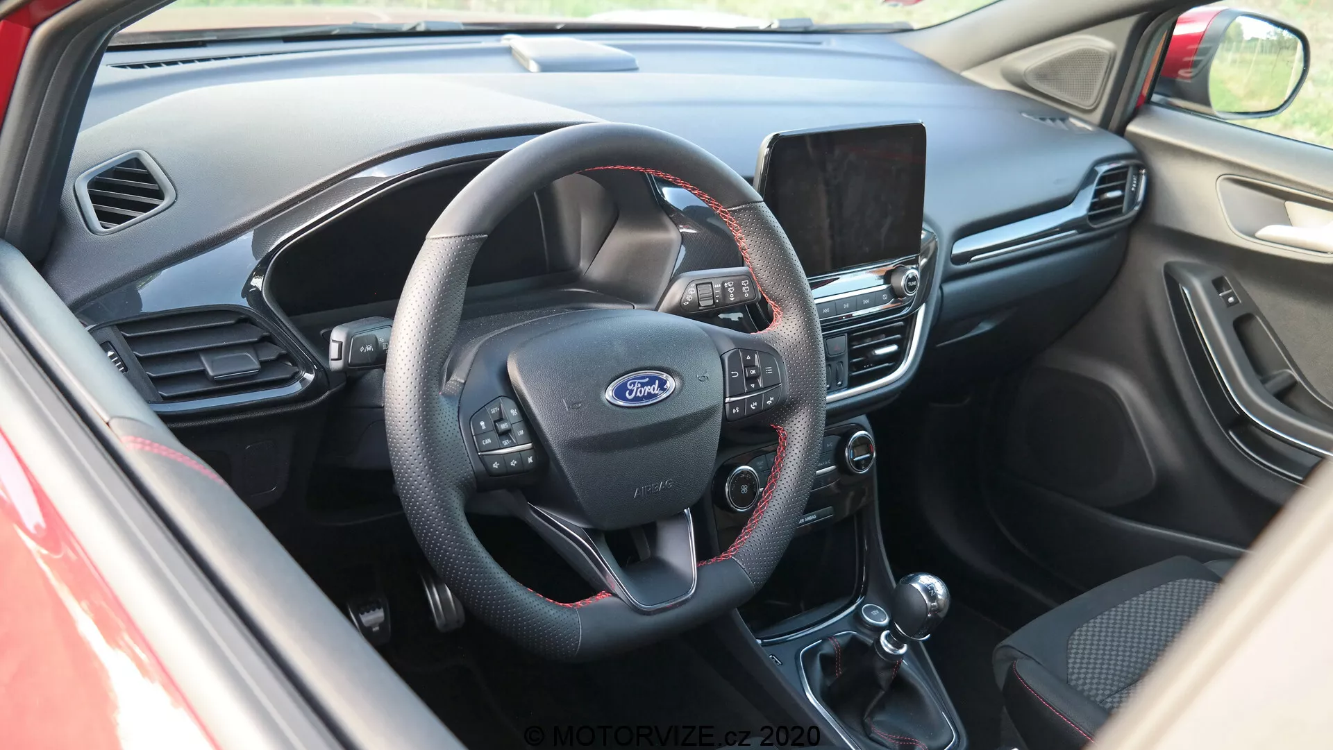 Vista interior del cockpit del Ford Puma 2020 desde un ángulo ligeramente elevado, enfocándose en el centro del tablero. Visto desde la puerta del conductor, la imagen muestra un volante de tres radios con botones multifuncionales para audio, comandos de voz y control de crucero. El emblema de Ford se muestra claramente en el centro. Detrás del volante se puede ver una parte del cuadro de instrumentos. El tablero central tiene una pantalla de infoentretenimiento que probablemente ofrece navegación, medios y configuraciones. Debajo, se encuentra el sistema de control de clima manual con diales giratorios y botones para temperatura, aire acondicionado y descongelación. La imagen también revela la palanca de cambios de transmisión automática y la manija del freno de mano. El interior combina plástico oscuro en la parte superior del tablero con tela o cuero negro en las partes inferiores, presentando un diseño funcional enfocado en la usabilidad.