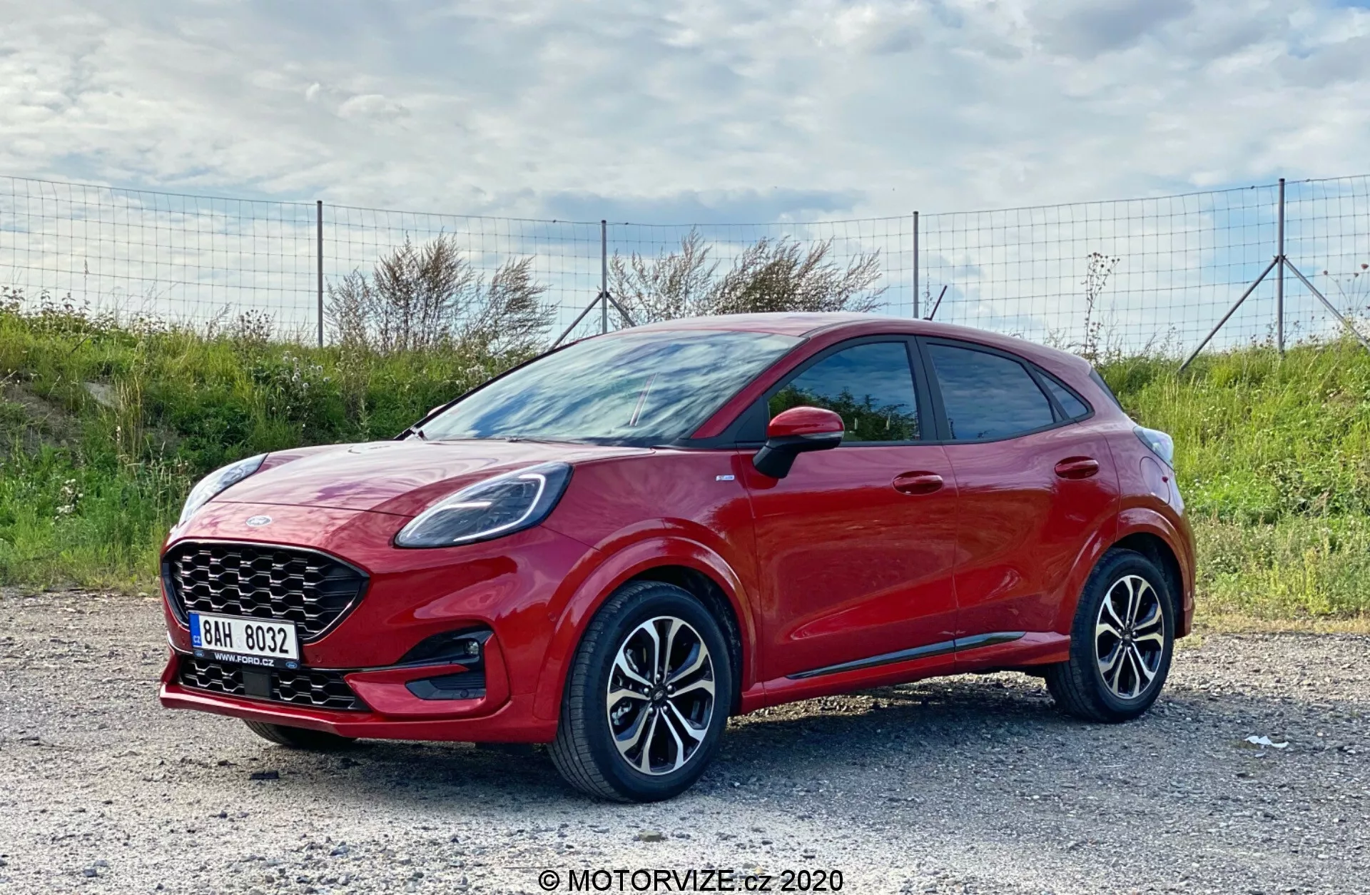 Imagen de perfil lateral de un hatchback moderno Ford Puma 2020, capturada a la luz del día con un fondo natural al aire libre. El vehículo de color rojo vibrante, con un acabado brillante, se ve desde un ángulo de tres cuartos del frente, mostrando claramente su perfil lateral y la fascia frontal. El coche está aparcado sobre una superficie de grava. La toma, realizada desde ligeramente arriba del nivel de las ruedas, resalta las llantas de aleación con diseño de múltiples radios y la parte inferior del vehículo. Son visibles los faros elegantes con probable tecnología LED, una parrilla frontal pronunciada y espejos laterales con indicadores integrados. El coche presenta manijas de puertas del color de la carrocería, sin molduras protectoras laterales visibles y un estilo deportivo hatchback con una línea del techo descendente. Las características tecnológicas posiblemente incluyen sistemas avanzados de asistencia al conductor y entretenimiento con opciones de conectividad.