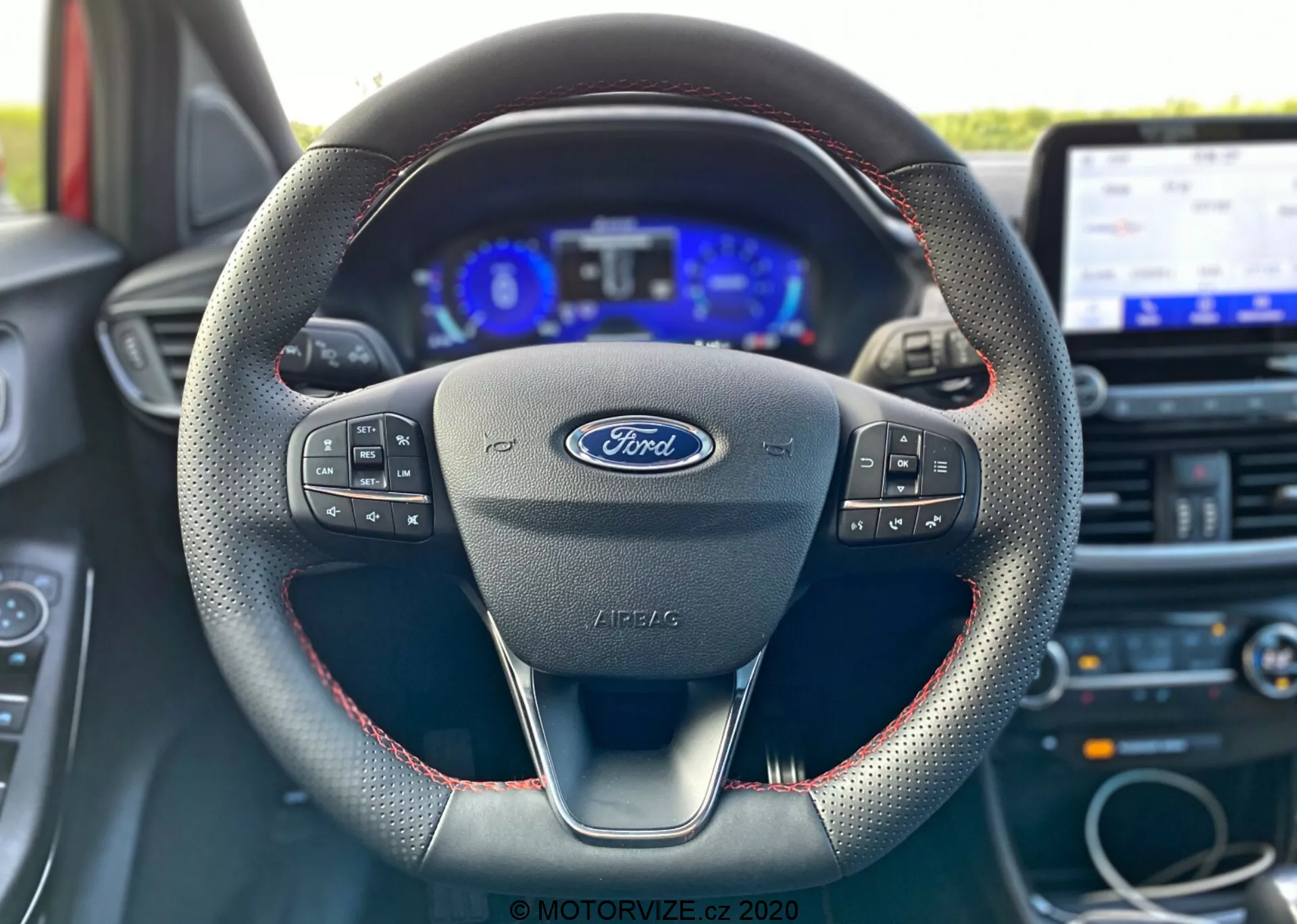 Imagen interior de un Ford Puma 2020 desde la perspectiva del conductor, enfocándose en el volante con el logo de Ford y controles multifuncionales, diseño moderno de tres radios con costuras rojas sugiriendo material de cuero, un cuadro de instrumentos digital flanqueado por indicadores analógicos y un sistema de infoentretenimiento con pantalla táctil en el fondo.