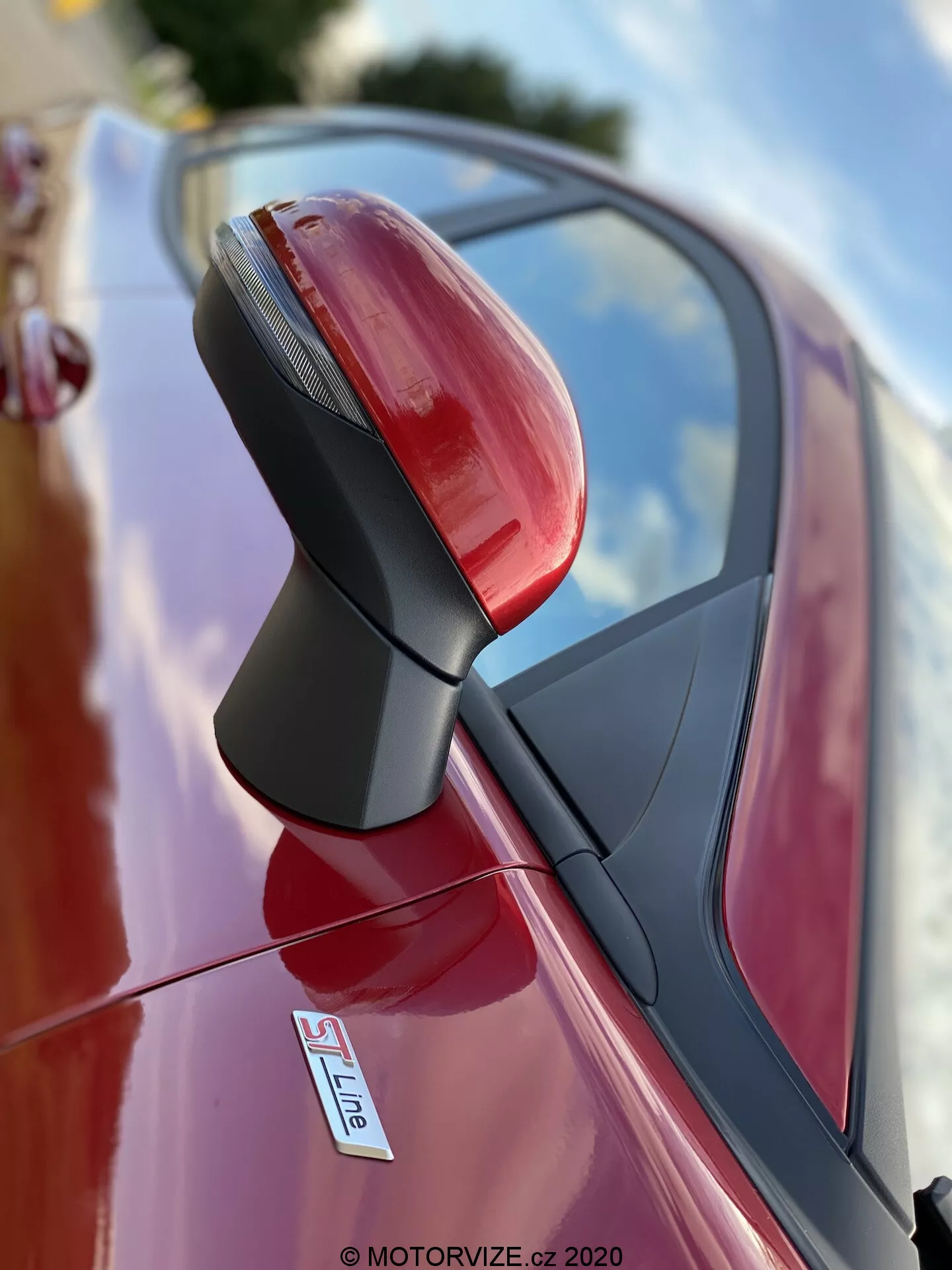 Primer plano del retrovisor exterior del lado del conductor de un coche rojo, probablemente un Audi con insignia S Line. La fotografía, tomada desde un ángulo bajo, se centra en el conjunto del espejo con una profundidad de campo reducida, difuminando el fondo. El espejo es nítido y detallado, mostrando señales de giro integradas, un soporte negro texturizado y un acabado metálico brillante. Los reflejos del cielo y el entorno indican que el coche está estacionado afuera.