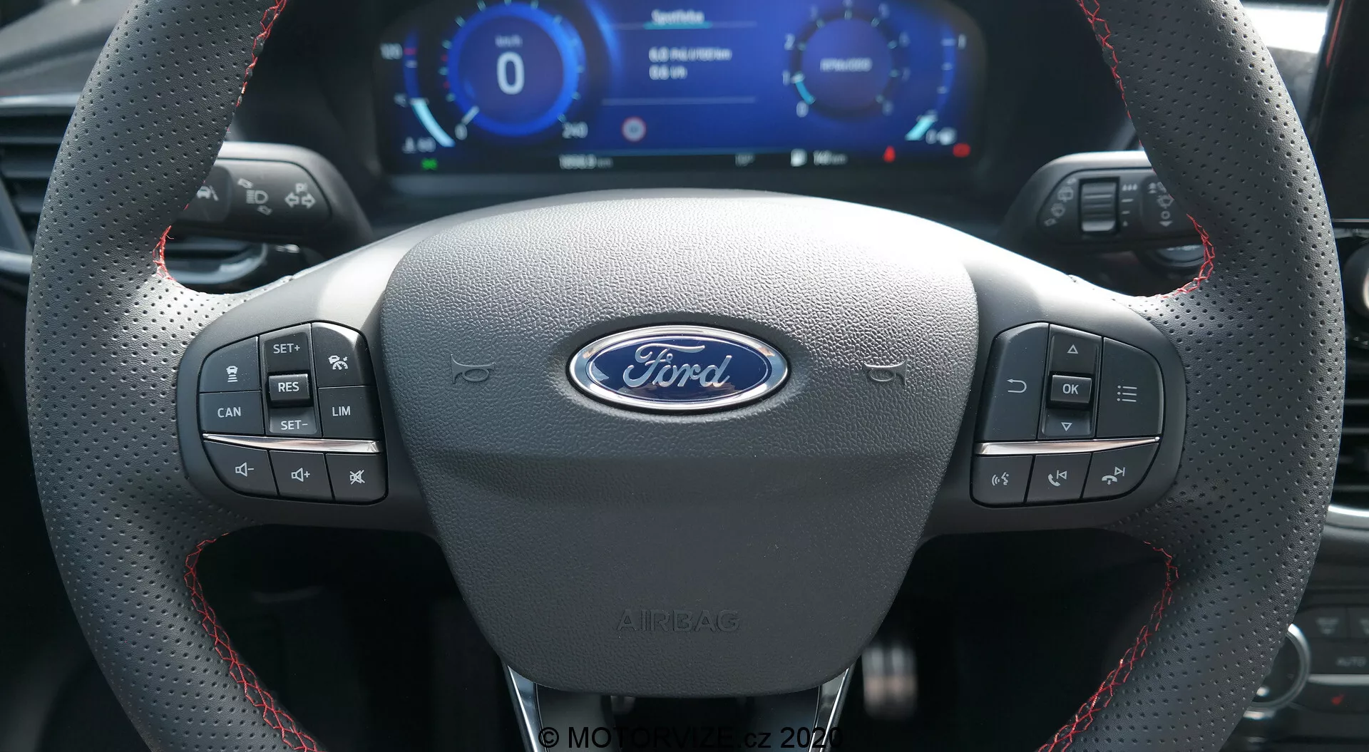 El interior de un Ford Puma 2020 desde la perspectiva del conductor que muestra un volante de cuero deportivo con costuras rojas, el logo central de Ford, múltiples botones de control, un cuadro de instrumentos digital con velocímetro y cuentarrevoluciones, y paletas de cambio metálicas.
