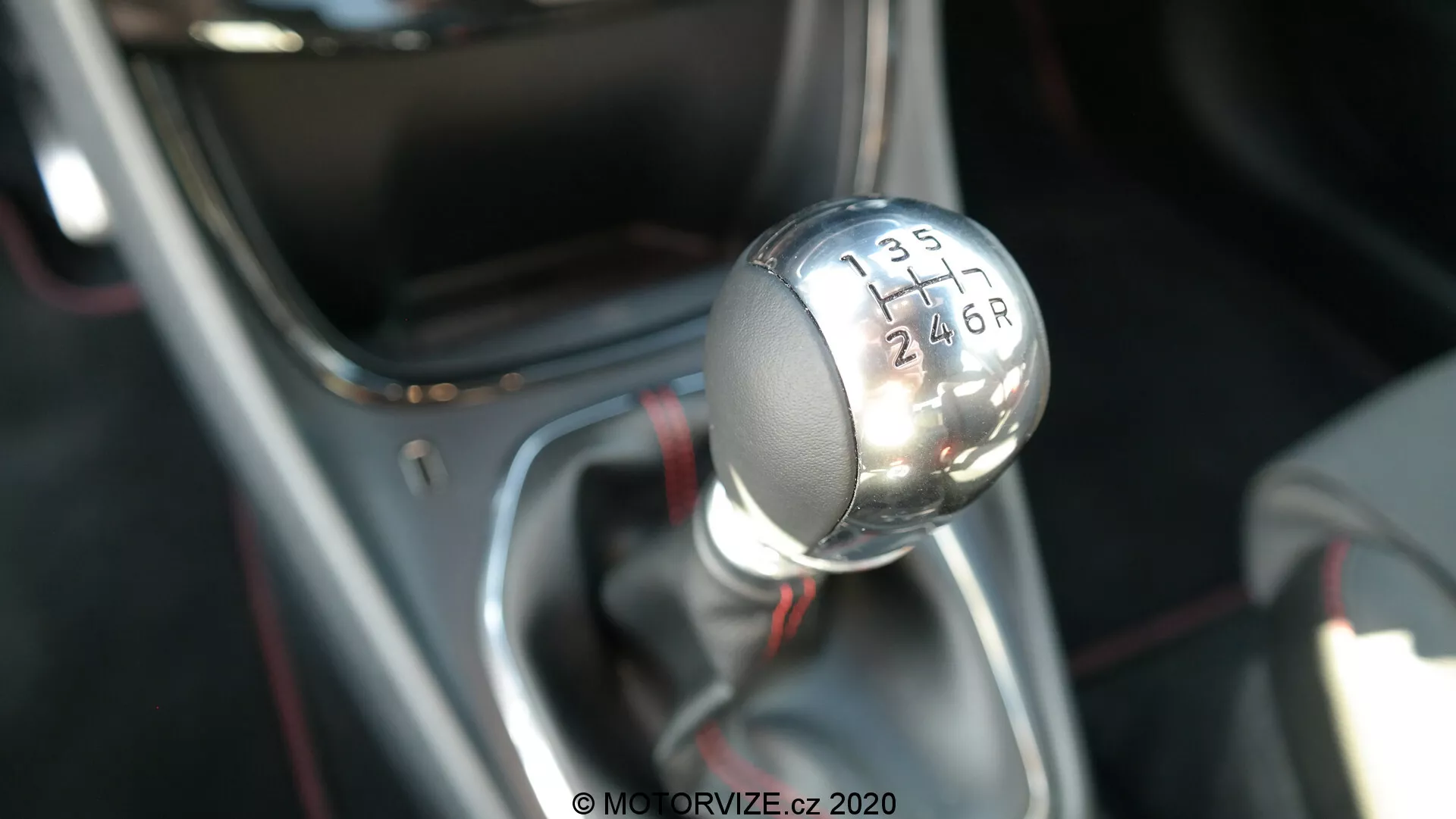 Fotografía en primer plano de la palanca de cambios manual del Ford Puma 2020, vista desde un ángulo ligeramente elevado que muestra el perfil superior y lateral. La perilla de cambio metálica tiene una parte superior negra con números blancos grabados que indican un patrón de transmisión de 6 velocidades con una 'R' para reversa. La palanca de cambios está montada en una base adornada de cromo y cubierta por una funda negra, enfocándose nítidamente en la perilla de cambios mientras que el fondo está desenfocado, destacando el interior y la tecnología del tren motriz del vehículo.