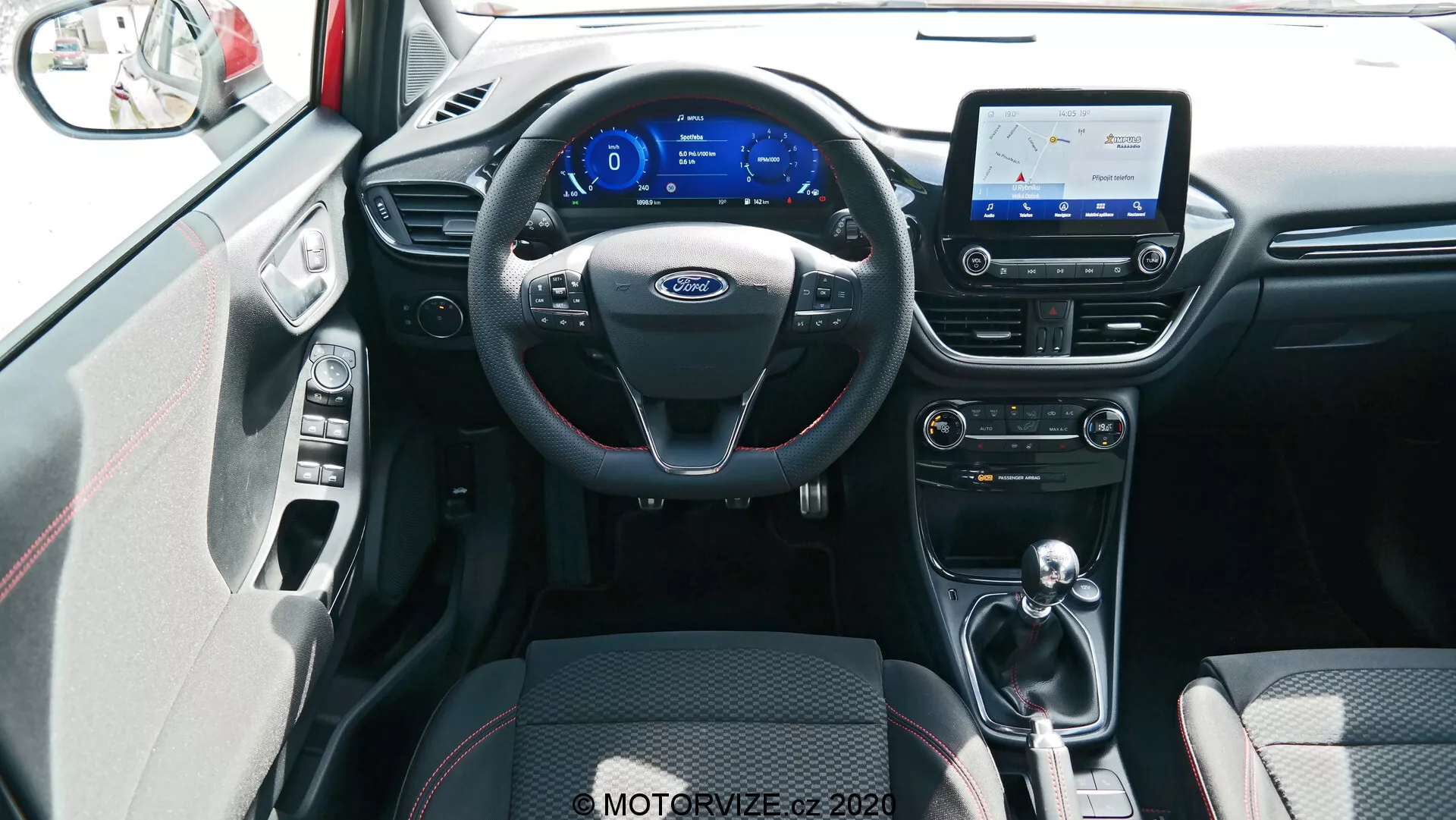 Vista interior del coche Ford Puma 2020, tomada desde la posición central del asiento trasero. La imagen muestra un volante de tres radios negro con controles multifuncionales y detalles plateados, un tablero digital con una pantalla de tema azul, y una pantalla táctil de infoentretenimiento que muestra un mapa, indicando capacidades de navegación. Debajo de la pantalla, hay controles para el aire acondicionado y la ventilación con diales y botones para ajustes de temperatura y velocidad del ventilador. El túnel central alberga una palanca de cambios, lo que sugiere una transmisión automática o manual automatizada. Los asientos delanteros presentan una tapicería de tela de dos tonos en negro con patrones para agarre y comodidad, y se ve una sección del panel interior de la puerta, incluyendo una manija de puerta plateada y detalles a juego. El suelo está cubierto de alfombras que combinan con el esquema de color interior y posiblemente hay buena iluminación ambiental. La toma se realiza desde un ángulo ligeramente elevado, mostrando de manera exhaustiva el interior delantero y las características tecnológicas disponibles.