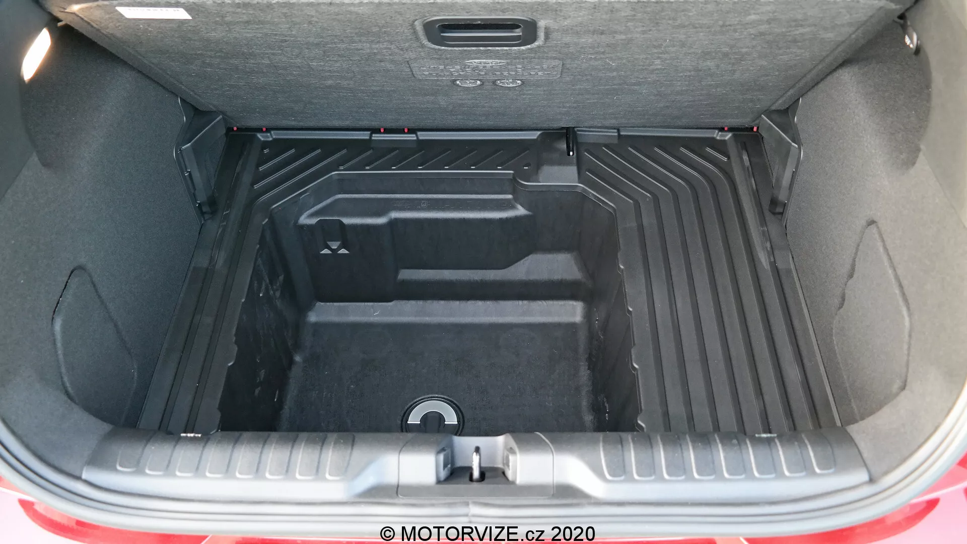 Una vista detallada del área espaciosa y versátil del maletero del Ford Puma 2020 con la tapa del maletero abierta, mostrando el interior vacío, limpio y cuidadosamente contorneado diseñado para la organización de equipaje y carga. El interior del maletero está hecho de un material duradero y fácil de limpiar, con secciones elevadas y hundidas para soluciones de almacenamiento optimizadas, incluido un asa empotrada central para acceder a un compartimiento inferior o al almacenamiento de la rueda de repuesto. Son parcialmente visibles el parachoques trasero y una franja del color rojo del exterior del coche, junto con la tapa aislada del maletero que tiene un asa para facilitar su cierre.