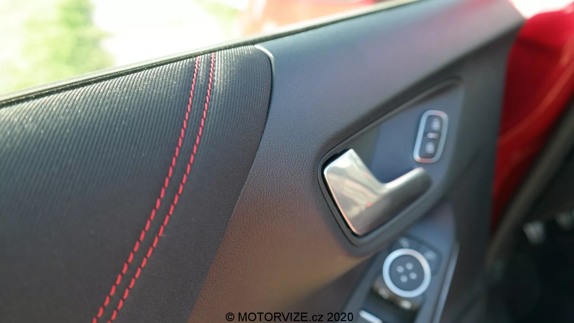 Vista en primer plano del panel interior de la puerta del Ford Puma 2020 desde la perspectiva de un pasajero o conductor dentro del vehículo. La imagen se enfoca en el área del tirador, destacando la inserción texturizada con costuras en contraste rojo, el tirador metálico de la puerta y parte del panel de controles con el interruptor de la ventana eléctrica. La poca profundidad de campo desenfoca el fondo, resaltando los elementos de diseño deportivos y de lujo bajo la luz natural del día.