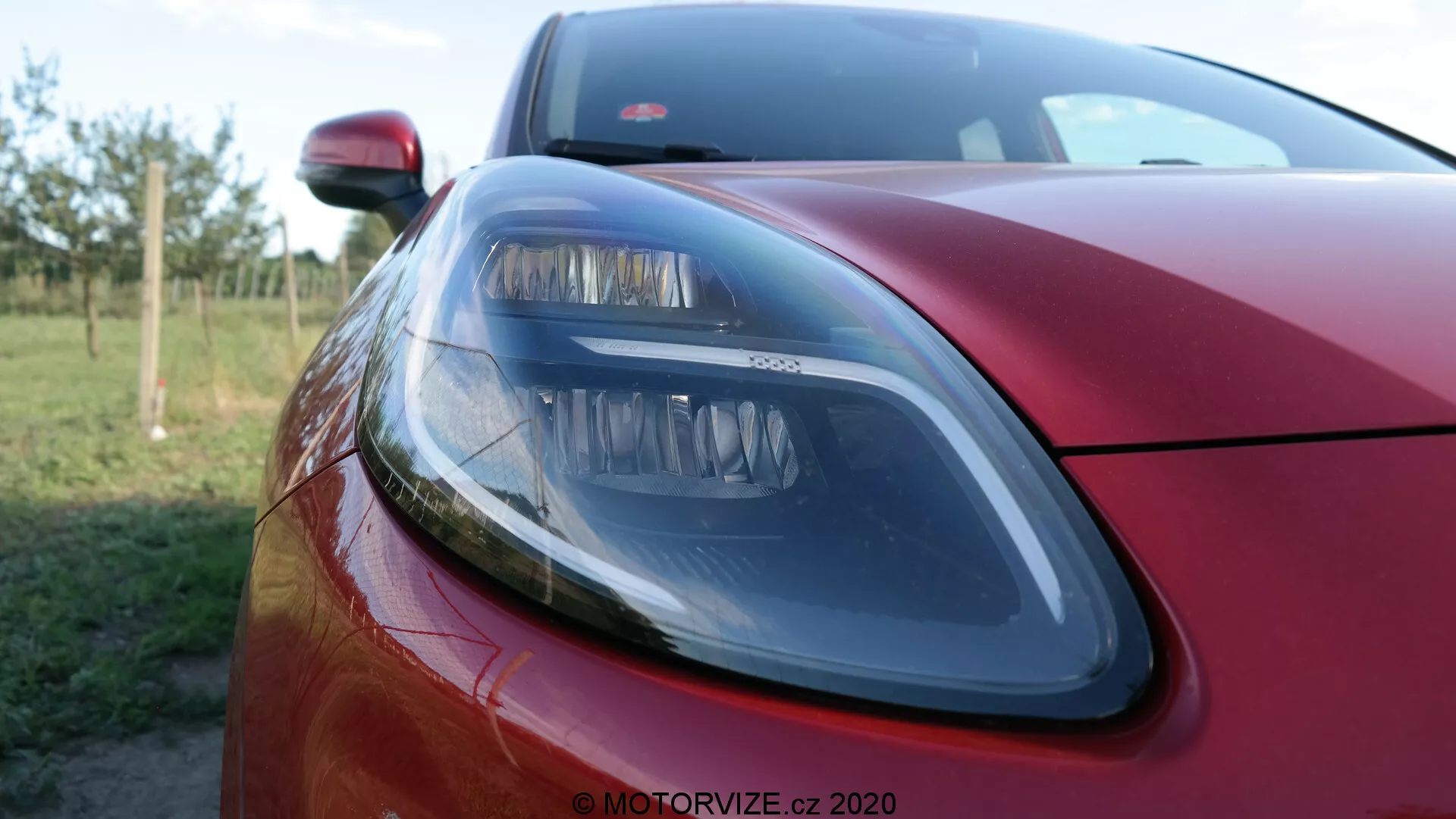 Primer plano de la sección delantera izquierda de un Ford Puma 2020 en un profundo rojo metálico, que muestra un faro moderno con elementos de diseño elegante, parte del capó y un vistazo a la parrilla delantera con una profundidad de campo reducida que desenfoca el fondo, destacando el acabado exterior premium y la tecnología de iluminación.