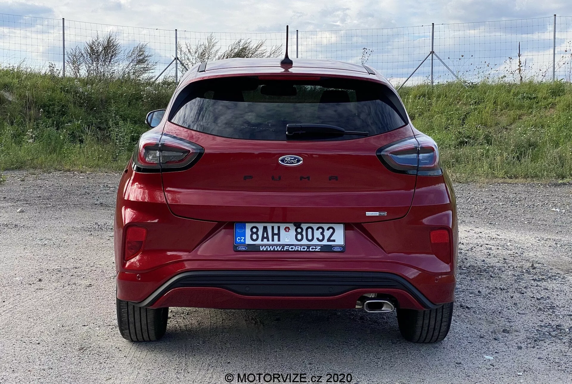 Vista trasera de un Ford Puma rojo 2020 hatchback sobre una superficie de grava, capturada en un ángulo nivelado y centrado. Las características incluyen un alerón trasero, limpiaparabrisas trasero, luz de freno alta, sistema de escape dual, superficies reflectantes, sensores de estacionamiento, cámara trasera y emblema Ford y nombre del modelo prominentes. Sin equipo de remolque o portaequipajes visible. Foto por MOTORVIZE.cz.