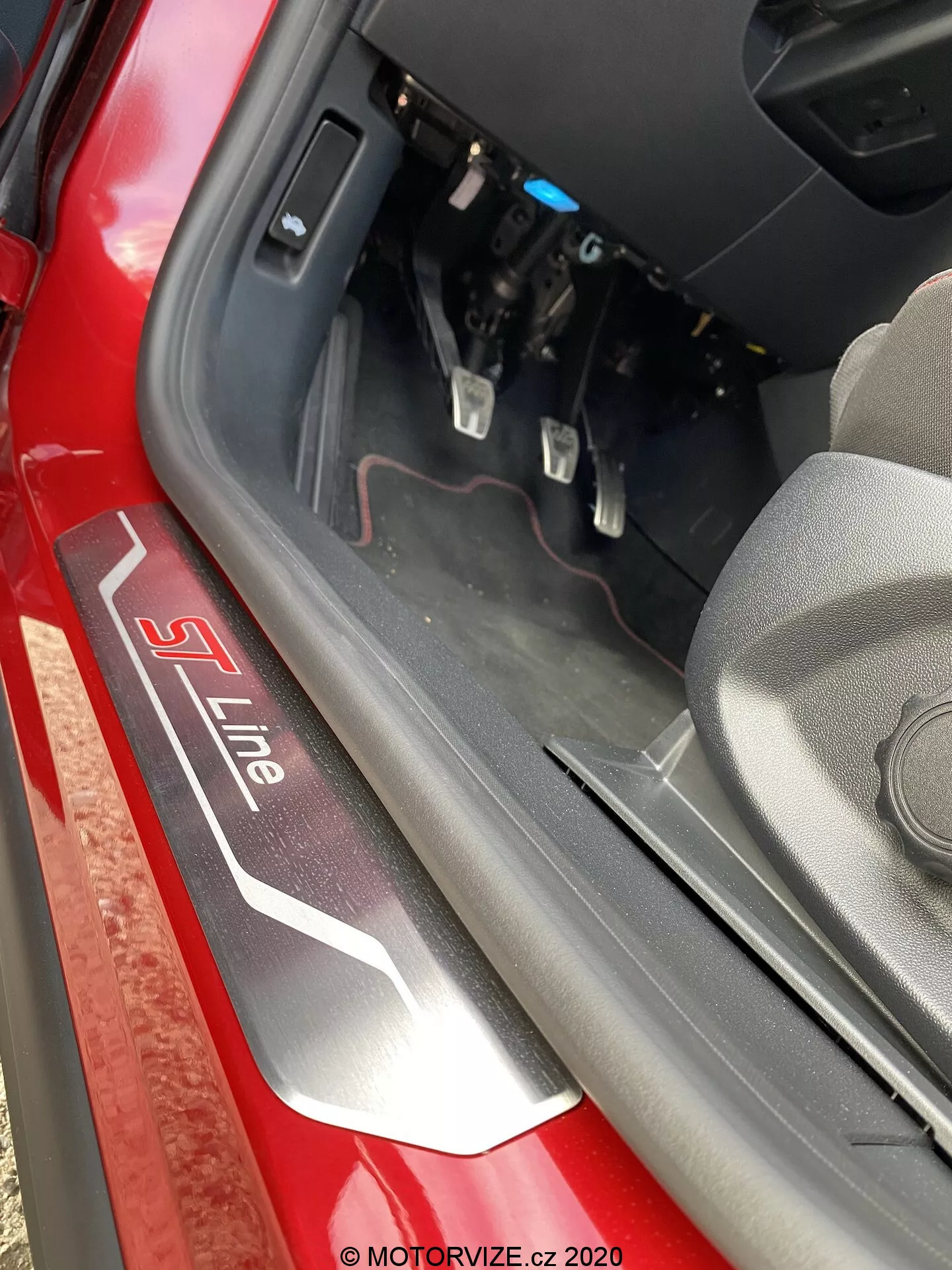 Vista de primer plano del umbral de la puerta de un vehículo Audi con la insignia 'S Line' en plata y rojo sobre un fondo negro. Tomado desde arriba y ligeramente hacia adentro, la imagen captura el borde inferior de la puerta, el borde interior y una porción del espacio para los pies del conductor con los pedales del freno y acelerador visibles. La luz natural del día ilumina la escena, destacando el color exterior rojo y los componentes interiores cerca del tablero.