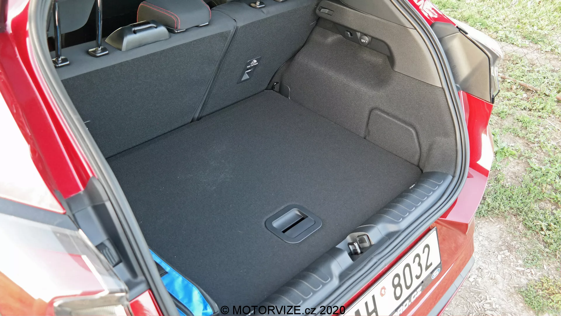 La imagen muestra el compartimento trasero o maletero de un Ford Puma 2020. La foto está tomada desde un ángulo ligeramente arriba y de lado, proporcionando una vista clara del espacioso interior del maletero y su umbral. La alfombra oscura sugiere que está diseñada para minimizar la apariencia de suciedad. No se ven elementos de carga, enfatizando la limpieza y el espacio del maletero. Las características visibles incluyen indentaciones laterales o bolsillos para almacenamiento de pequeños artículos y un gancho o punto de amarre para asegurar objetos. El grupo óptico trasero del coche es parcialmente visible en el lado derecho de la imagen.