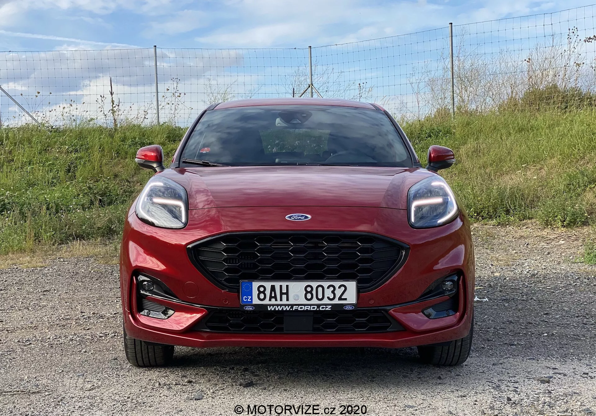 Ford Puma 2020 rojo capturado desde un ángulo frontal sobre una superficie de grava, mostrando la distintiva parrilla de Ford con el logo centrado, elegantes faros con luces diurnas LED, luces antiniebla integradas y matrícula centralmente montada. Ideal para el diseño del frente del vehículo, identificación de la marca y sistemas de iluminación automotriz modernos.