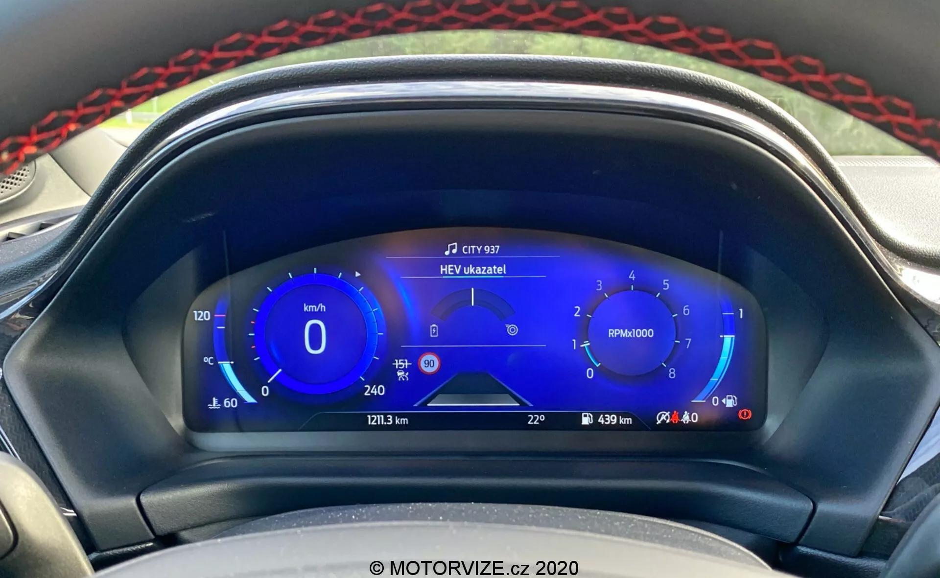 Vista de cerca del moderno tablero de instrumentos digitales del Ford Puma 2020 visto desde el asiento del conductor. La pantalla digital presenta un velocímetro con rangos de velocidad ecológicos a la izquierda, un tacómetro en el centro, medidores de combustible híbridos a la derecha y información adicional como kilometraje, distancia del viaje, alcance y temperatura exterior en la parte inferior. En la parte superior central aparece una notificación que dice 'HEV úzkazník!', indicando el modo de vehículo eléctrico híbrido. El diseño elegante y enfocado en la tecnología en tonos de azul, blanco y rojo resalta los avanzados sistemas de información para el conductor y la tecnología híbrida del vehículo.