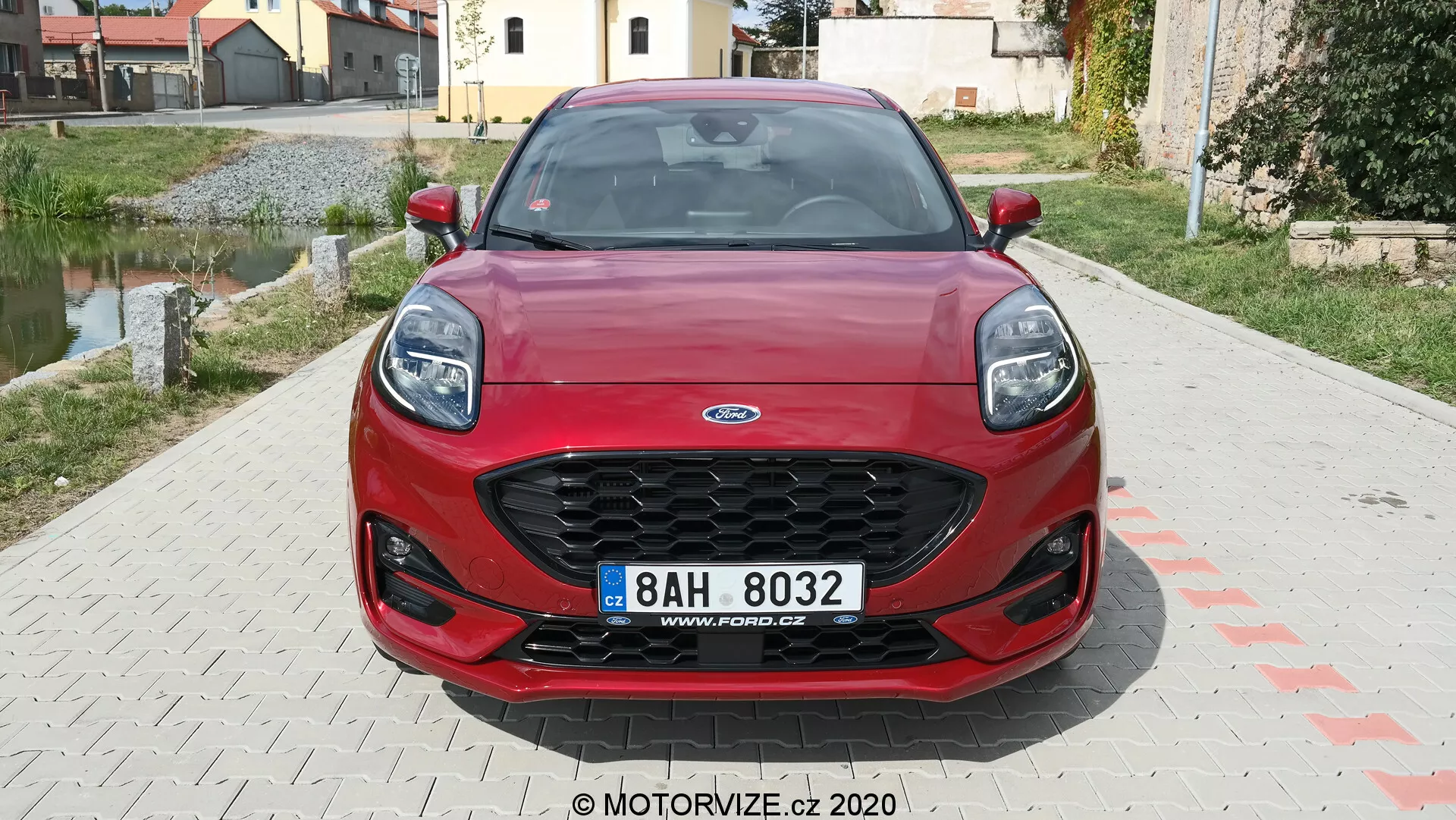 Vista frontal de un Ford Puma rojo 2020, que muestra una parrilla hexagonal con malla negra, faros modernos barridos hacia atrás, parachoques delantero deportivo con amplias tomas de aire y placa de matrícula central. Las líneas de contorno dinámicas del capó realzan la estética del vehículo. El fondo muestra una zona residencial.