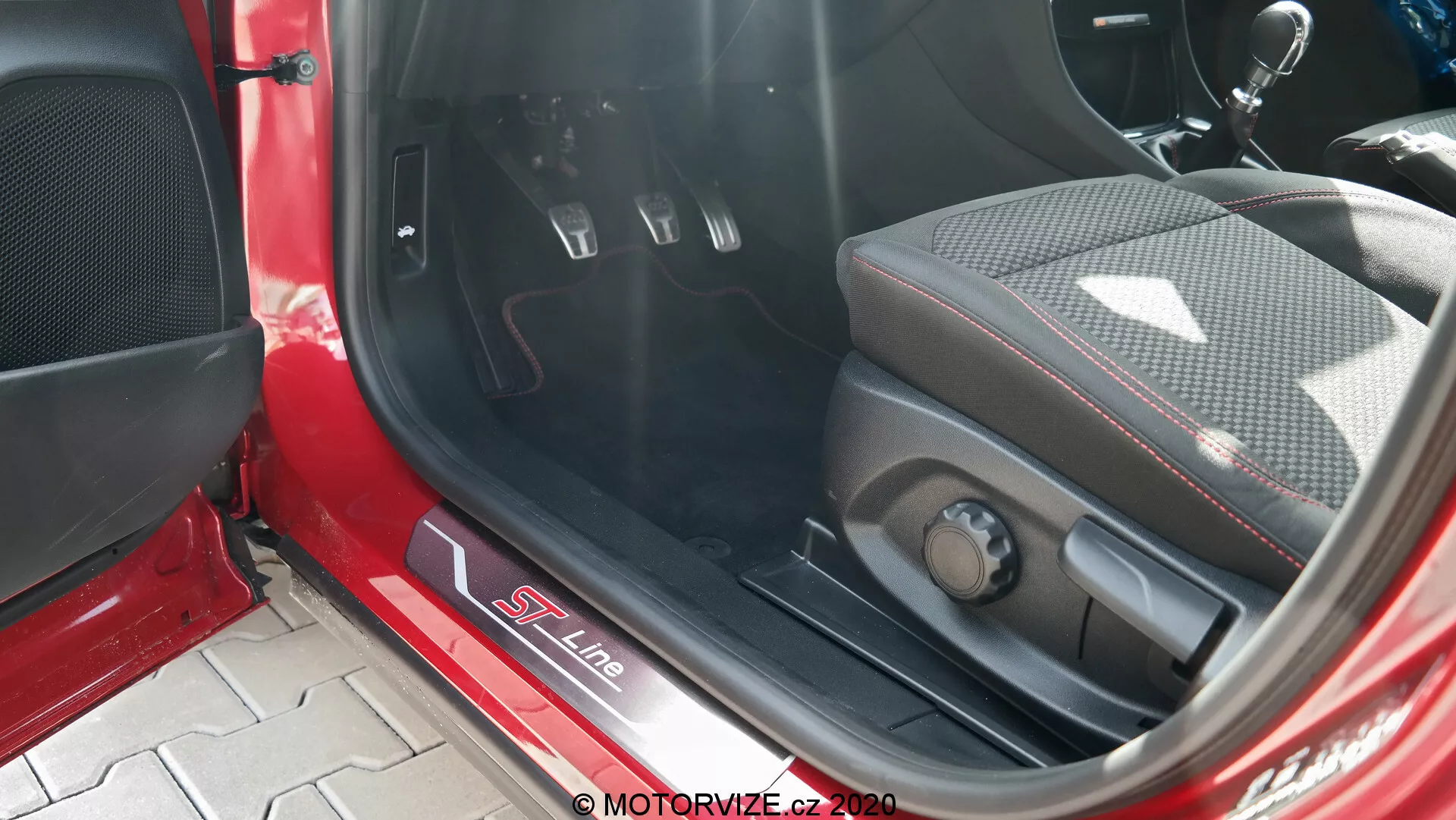Vista de cerca del interior de un Ford Puma 2020, enfocándose en el umbral de la puerta metálico con la marca S Line, asiento deportivo de mezcla de cuero y tela con refuerzos laterales, controles de ajuste del asiento electrónicos, pedales metálicos con inserciones de goma para transmisión manual, alfombrilla de piso alfombrada, panel interior rojo y negro con rejilla de altavoz y luz solar directa iluminando el interior.