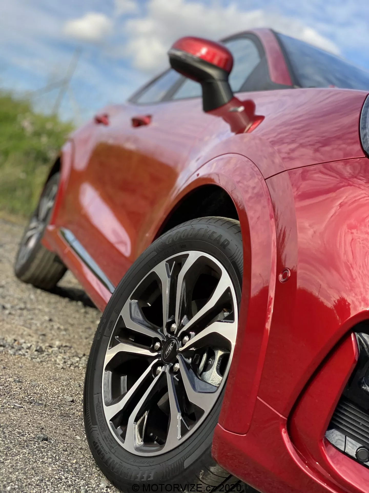 Vista de cerca de un coche deportivo Ford Puma 2020 rojo, enfocando en la rueda trasera y panel lateral. La toma a nivel del suelo desde un ángulo bajo resalta el diseño sofisticado de la llanta de múltiples radios, neumático de perfil bajo y el acabado de pintura brillante. La imagen captura intrincados detalles del diseño de la llanta y el patrón de la banda de rodadura, con una poca profundidad de campo que mantiene la rueda enfocada mientras el fondo se desdibuja. Se ve una pequeña entrada de aire detrás de la rueda en el panel lateral, destacando las características de rendimiento y aerodinámicas del vehículo.