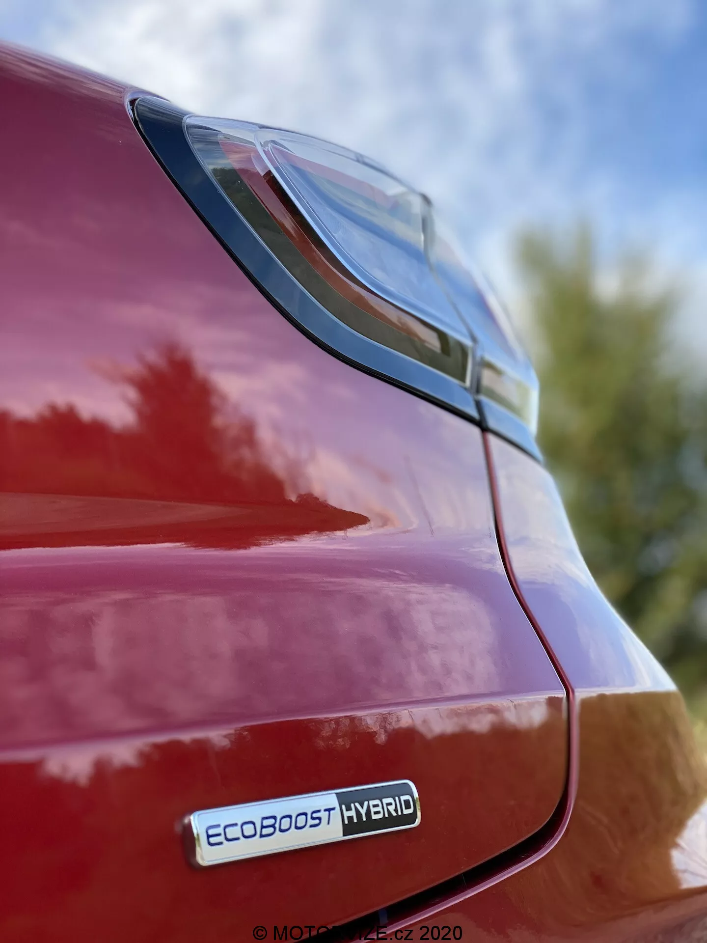 Vista de cerca de la parte trasera derecha de un Ford Puma rojo 2020, enfocándose en la luz trasera LED y el área del guardabarros trasero desde una perspectiva de ángulo bajo con la insignia EcoBoost Hybrid visible, reflejando un acabado brillante y un diseño elegante contra un fondo de cielo y follaje.