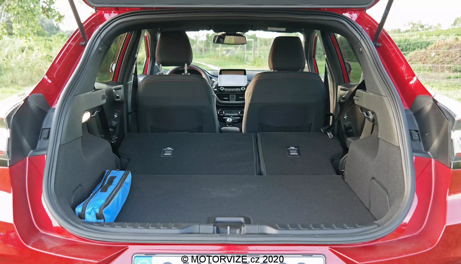 Imagen del área de carga del Ford Puma 2020 vista desde la parte trasera, mostrando el maletero abierto con los asientos traseros plegados, una bolsa azul a la derecha y una tapa retráctil vertical en el lado izquierdo, destacando el espacio de almacenamiento adaptable del hatchback