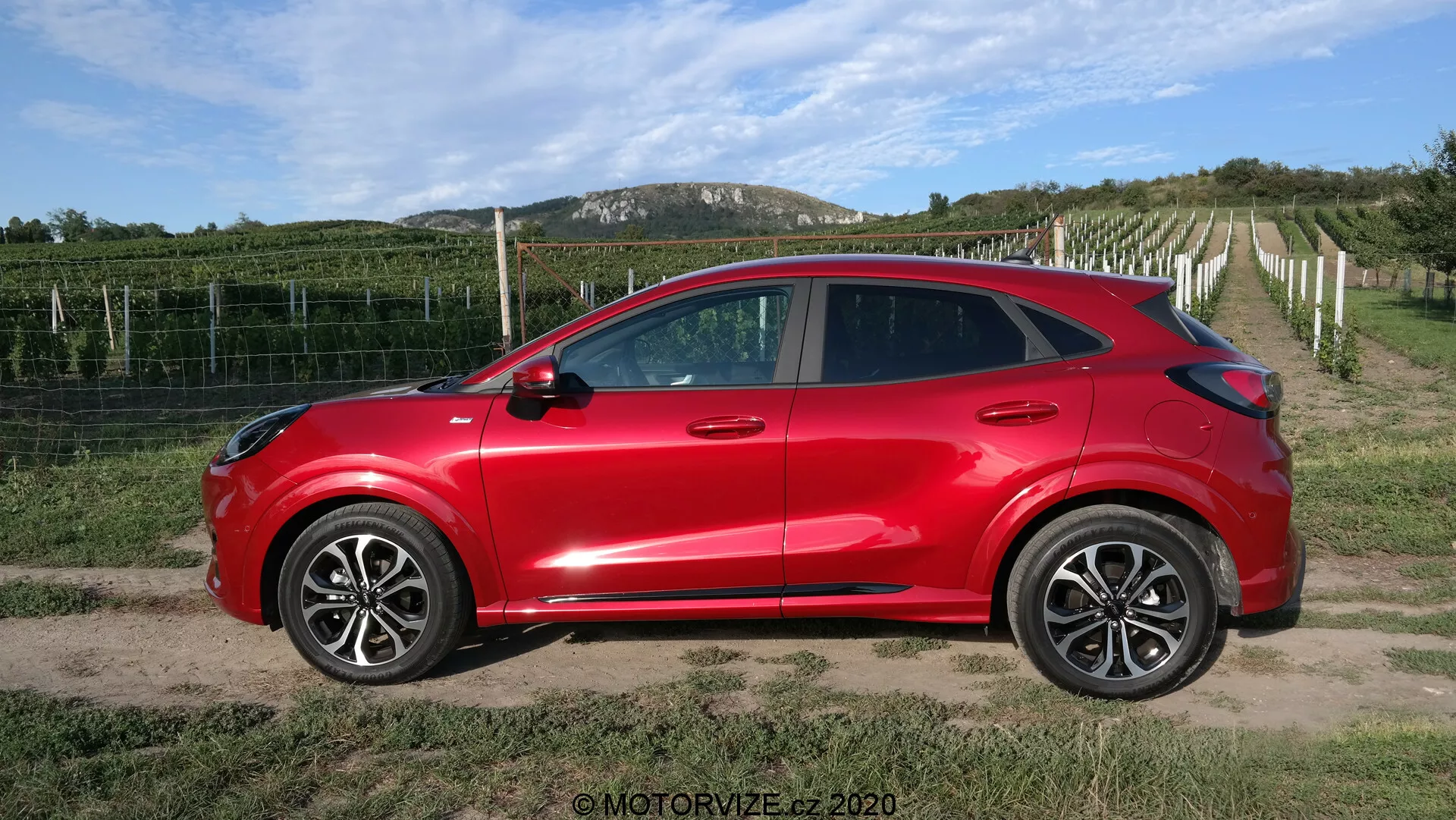 Vista lateral de un Ford Puma SUV crossover compacto de 2020 en color rojo brillante con cuatro puertas, grandes llantas de aleación, espejos laterales del color de la carrocería con indicadores de giro integrados, ventanas tintadas y manijas de las puertas del color de la carrocería. El vehículo, con su diseño aerodinámico moderno y línea de techo descendente, está situado en un camino de tierra o grava con un viñedo y cielo despejado en el fondo, proporcionando un ambiente rural.
