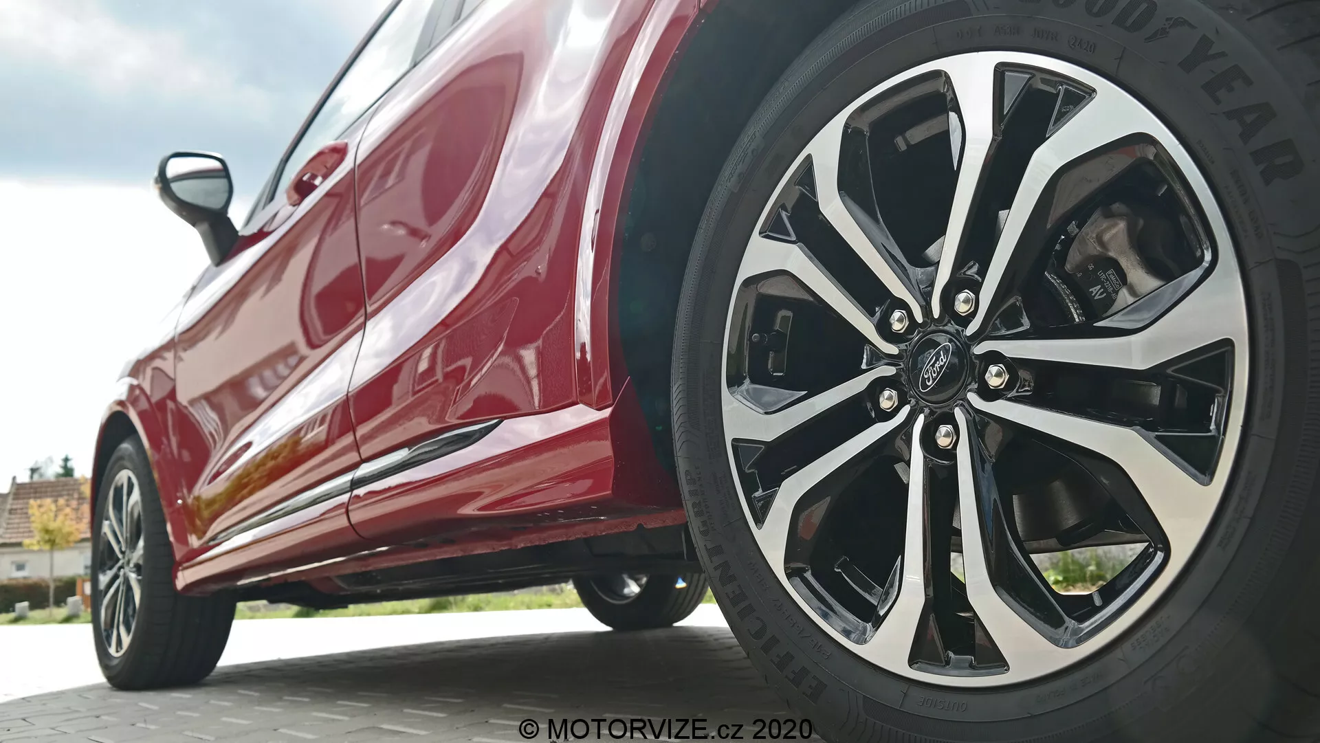 Vista cercana y a bajo ángulo del Ford Puma 2020 enfocándose en el perfil lateral cerca de la rueda delantera. La toma es desde el nivel del suelo, destacando el intrincado diseño de múltiples radios de la rueda del lado del conductor en una mezcla de acabados plateados o cromados y negros, junto con el neumático de marca Goodyear. La carrocería roja y brillante refleja la luz, mostrando elementos modernos como estribos laterales y líneas aerodinámicas.
