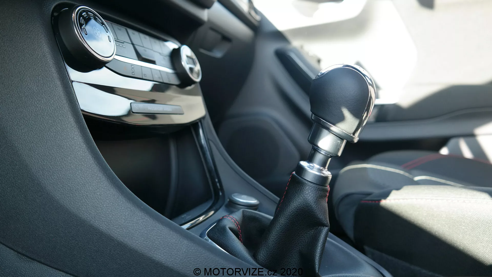Interior del coche Ford Puma 2020, centrado en la consola central desde la perspectiva del pasajero. La imagen destaca una palanca de cambios manual de seis velocidades en plástico oscuro con un embellecedor negro piano brillante alrededor de su base. En el fondo se vislumbra el volante parcialmente desenfocado, con botones de control de audio y crucero, junto con una salida de ventilación del tablero. La escena está iluminada por la luz natural del día.