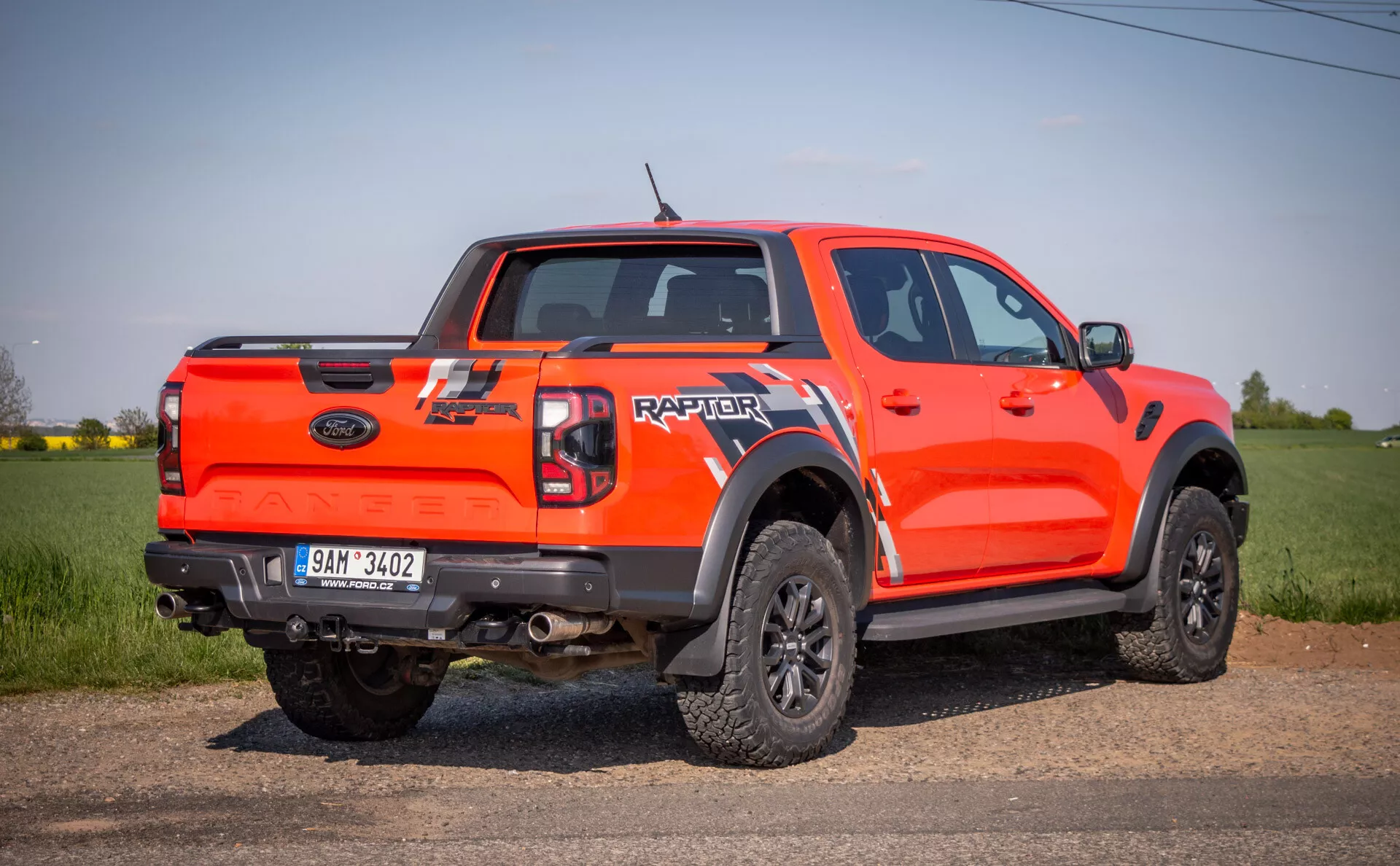 Camioneta Ford Raptor 2022 de color naranja brillante en perspectiva trasera-lateral, al aire libre en un campo, mostrando sus capacidades todoterreno con alta distancia al suelo, cabina doble, grandes extensiones de arco de rueda negras, ruedas negras de múltiples radios con neumáticos todoterreno, y el nombre 'RAPTOR' en grande en la puerta trasera.