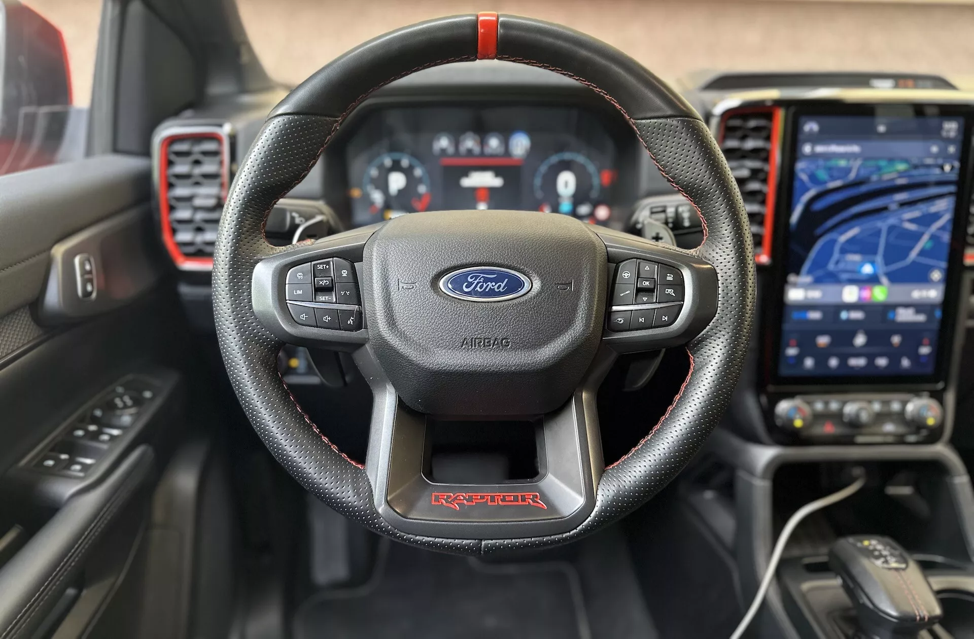 Vista frontal interior del Ford Ranger Raptor 2022 destacando el volante de tres radios envuelto en cuero con costuras en rojo y logo de Ford, cluster de instrumentos digital, sistema de infotenimiento táctil moderno en modo de navegación, tablero deportivo con acentos rojos y ayudas avanzadas para el conductor para una experiencia de conducción enfocada en la calidad y tecnología