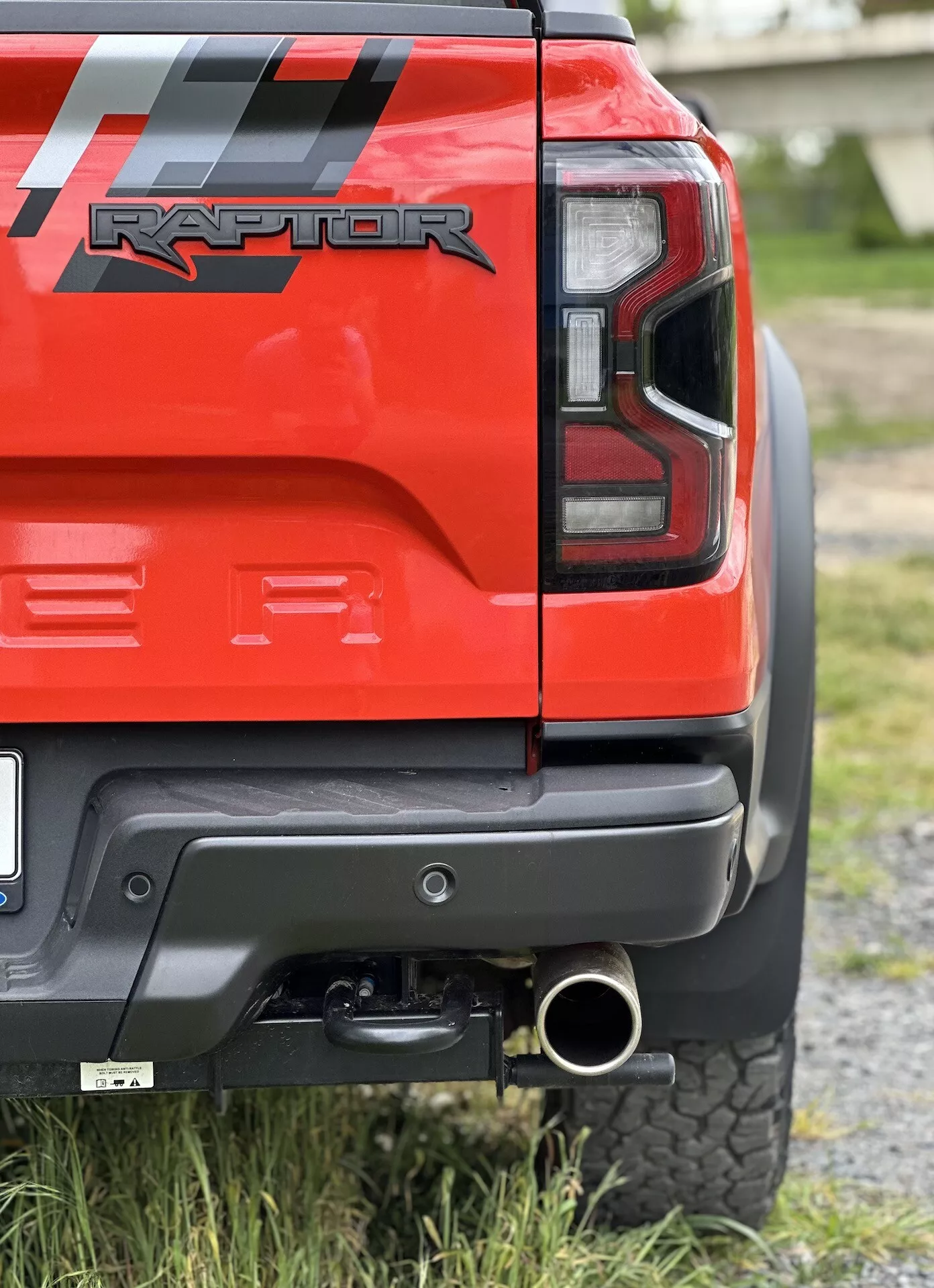 Vista cercana de la esquina trasera de un Ford Ranger Raptor rojo 2022 mostrando la luz trasera LED, parachoques trasero negro con panel gris, enganche de remolque, punta de escape pulida y neumático todoterreno, enfatizando sus capacidades fuera de carretera y detalles de diseño.