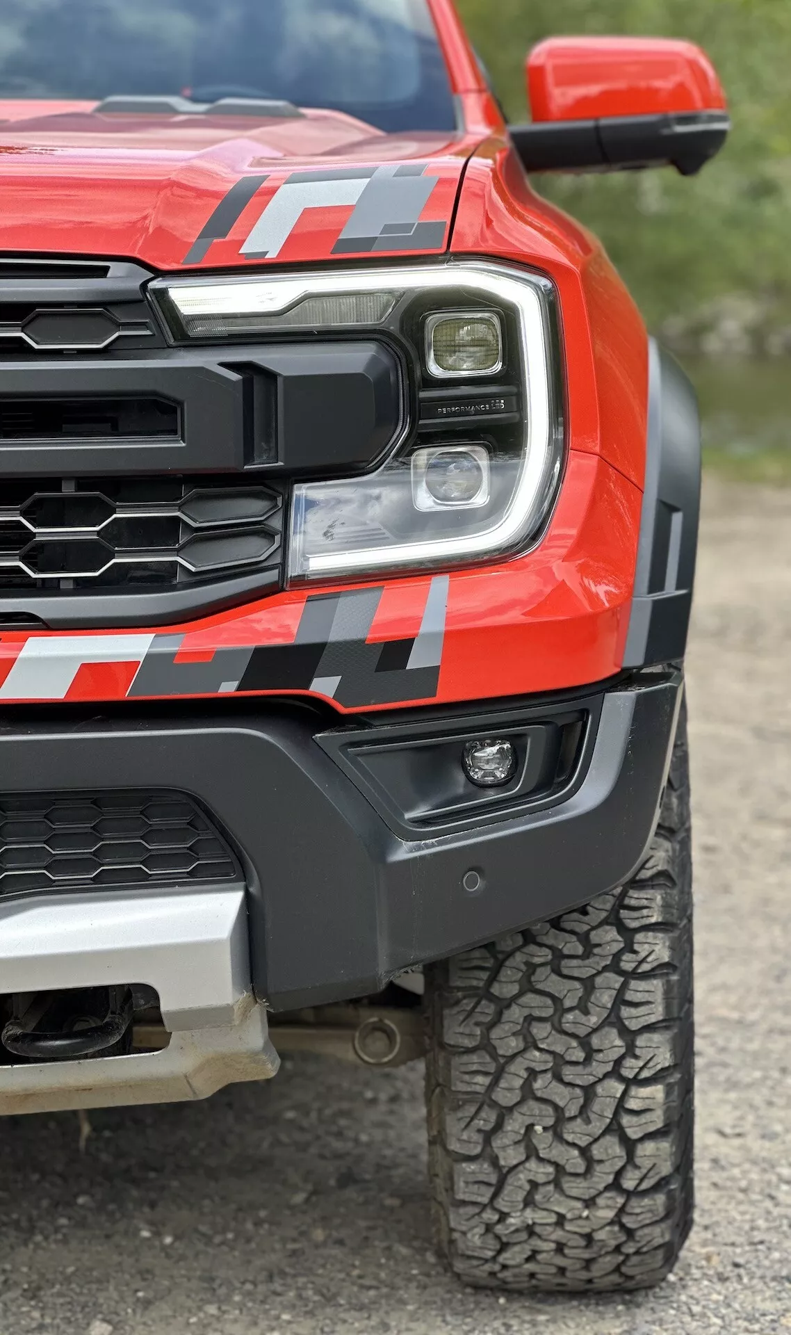 Vista cercana de una camioneta Ford Ranger 2022 en rojo, mostrando el faro LED, la rejilla robusta, el guardabarros frontal musculoso con calcomanía y el patrón de banda de rodadura de los neumáticos para todo terreno desde un ángulo bajo para enfatizar la robustez y tecnología avanzada