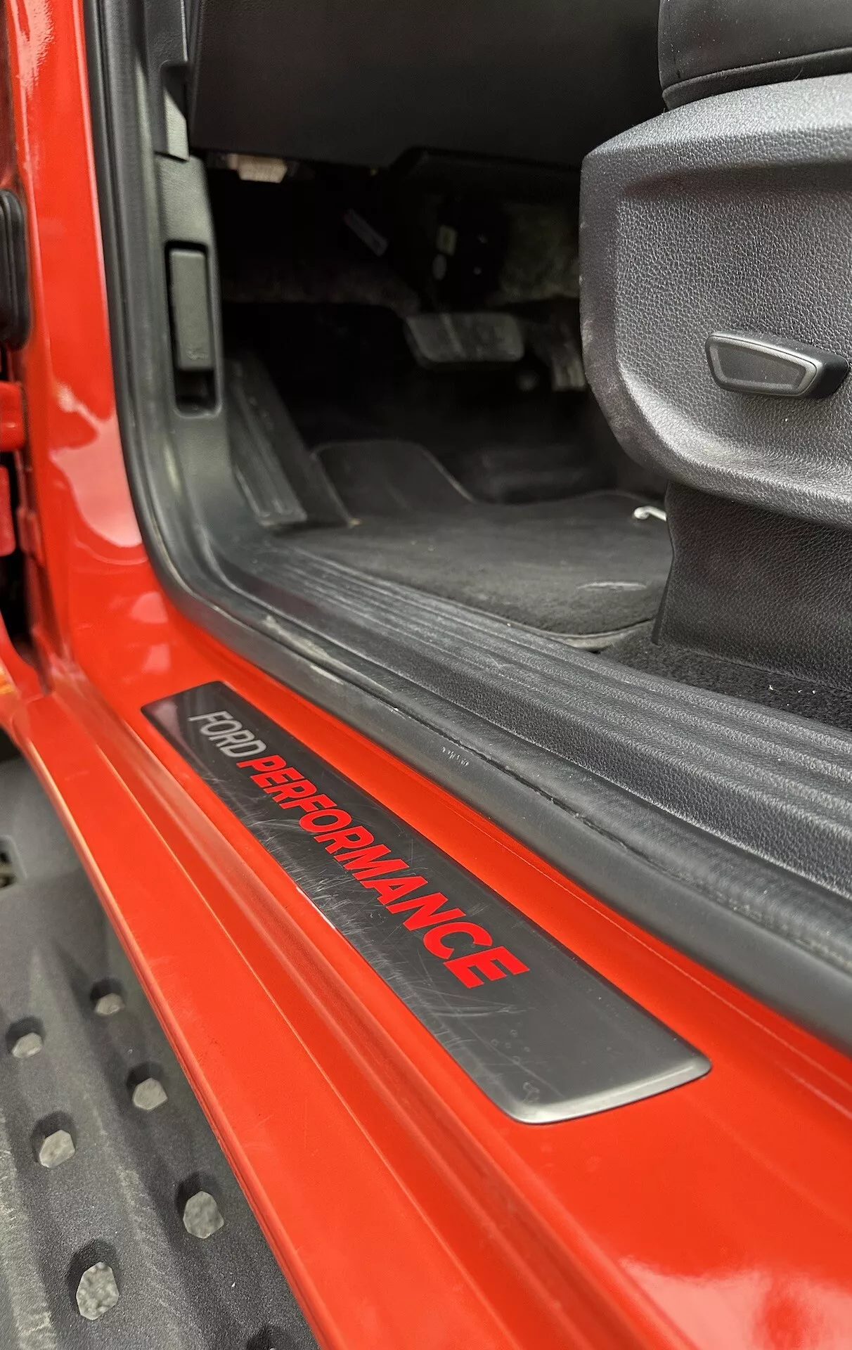 Primer plano del umbral de la puerta del Ford Ranger 2022 mostrando la insignia 'FORD PERFORMANCE', exterior rojo brillante, alfombra interior negra, asiento de tela gris con diseño deportivo y alfombrilla de goma negra duradera