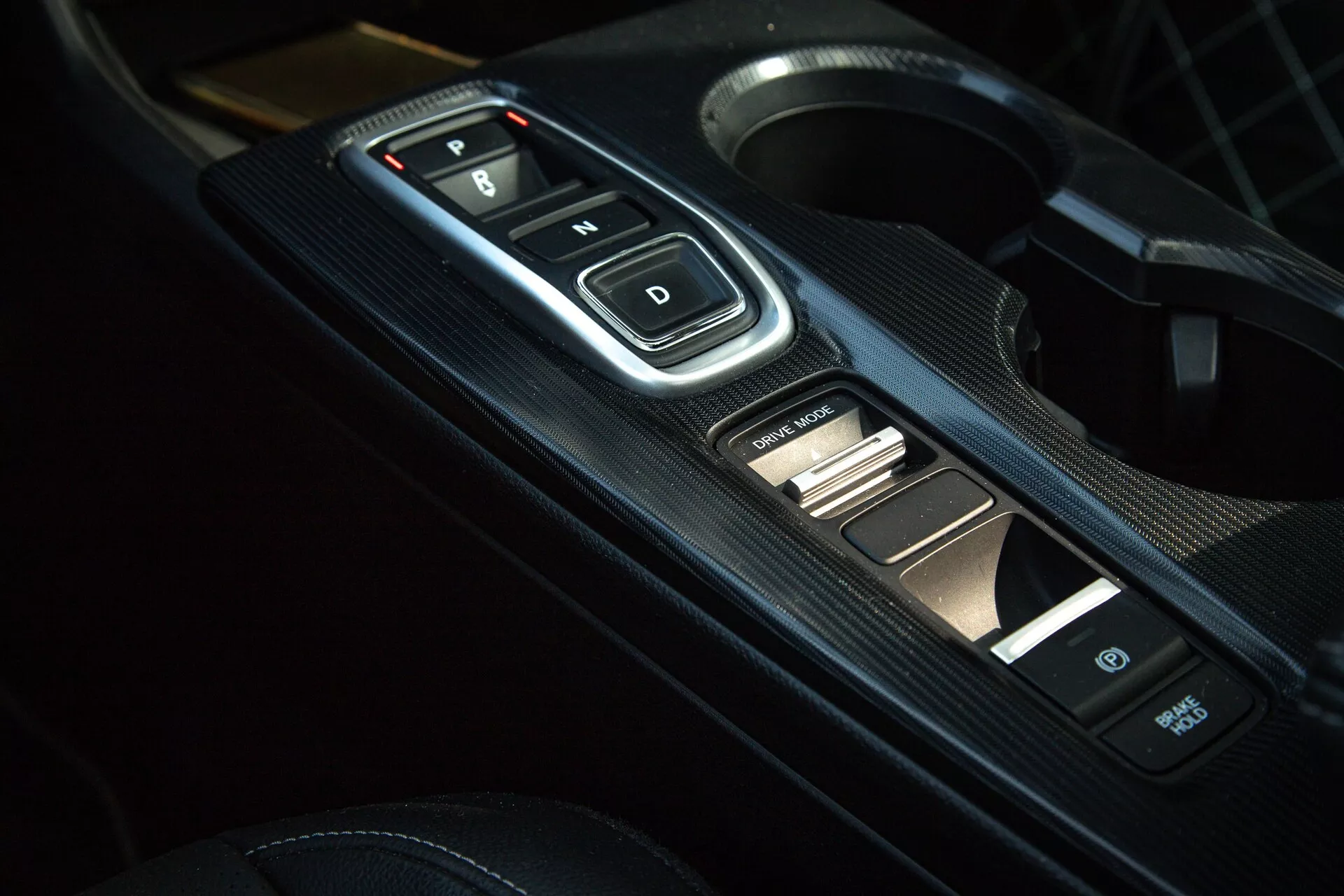 Primer plano del interior del Honda Civic 2022 de la consola central con el selector de marchas electrónico con botones 'P, R, N, D', panel con textura de fibra de carbono y controles auxiliares, mostrando la transmisión automática moderna y estética deportiva