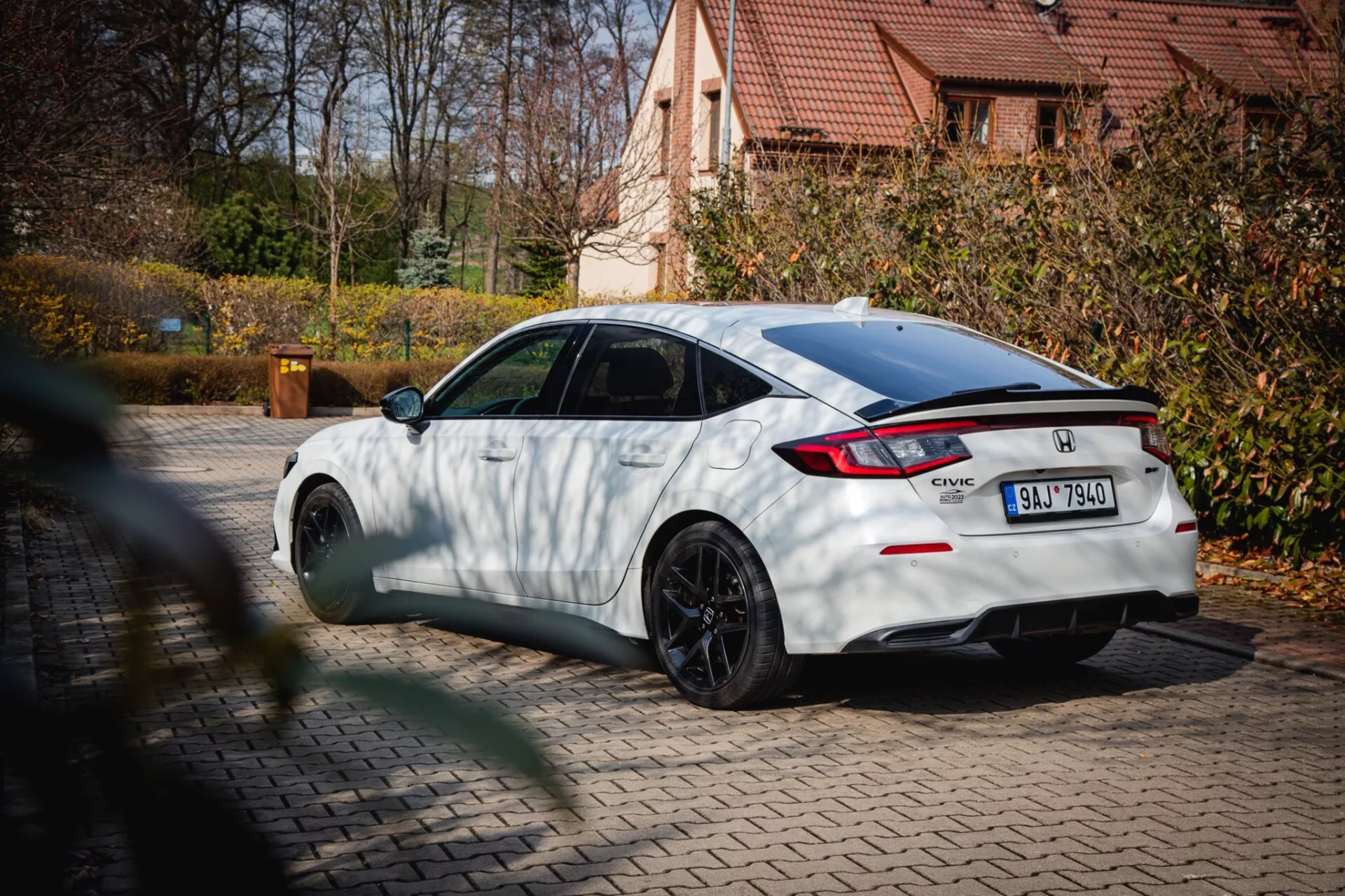 Honda Civic blanco del 2022 con ruedas multirradio negras y spoiler trasero, estacionado en un pavimento con follaje otoñal y una casa residencial al fondo