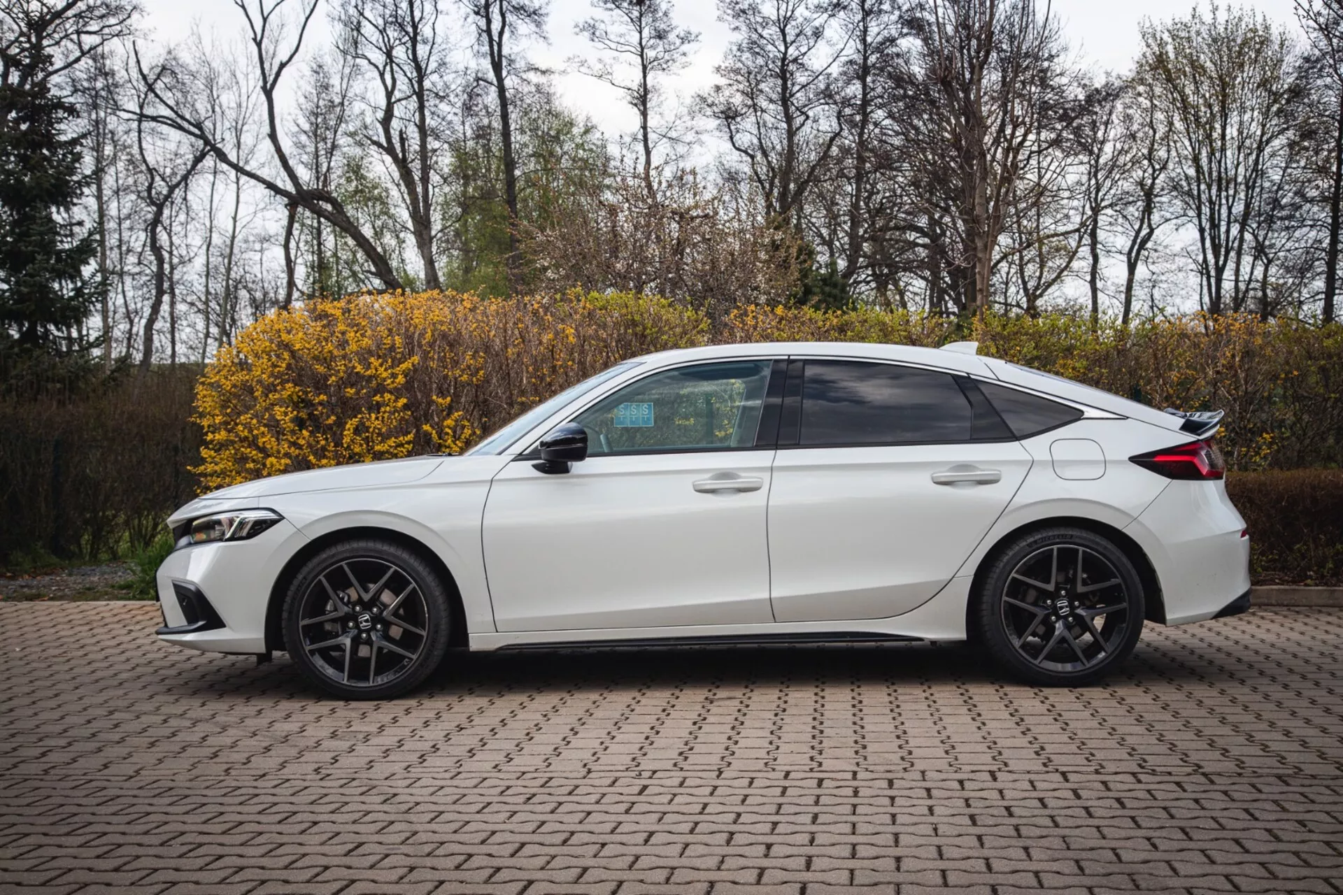 Sedán Honda Civic blanco de cuatro puertas y modelo 2022 estacionado sobre un área pavimentada con ladrillos entrelazados, mostrando un perfil lateral con un diseño deportivo y faros modernos LED, capturado desde un ángulo ligeramente elevado para resaltar la vista lateral completa incluyendo ventanas tintadas, llantas de aleación deportivas y características tecnológicas como señales de giro integradas en los espejos laterales.