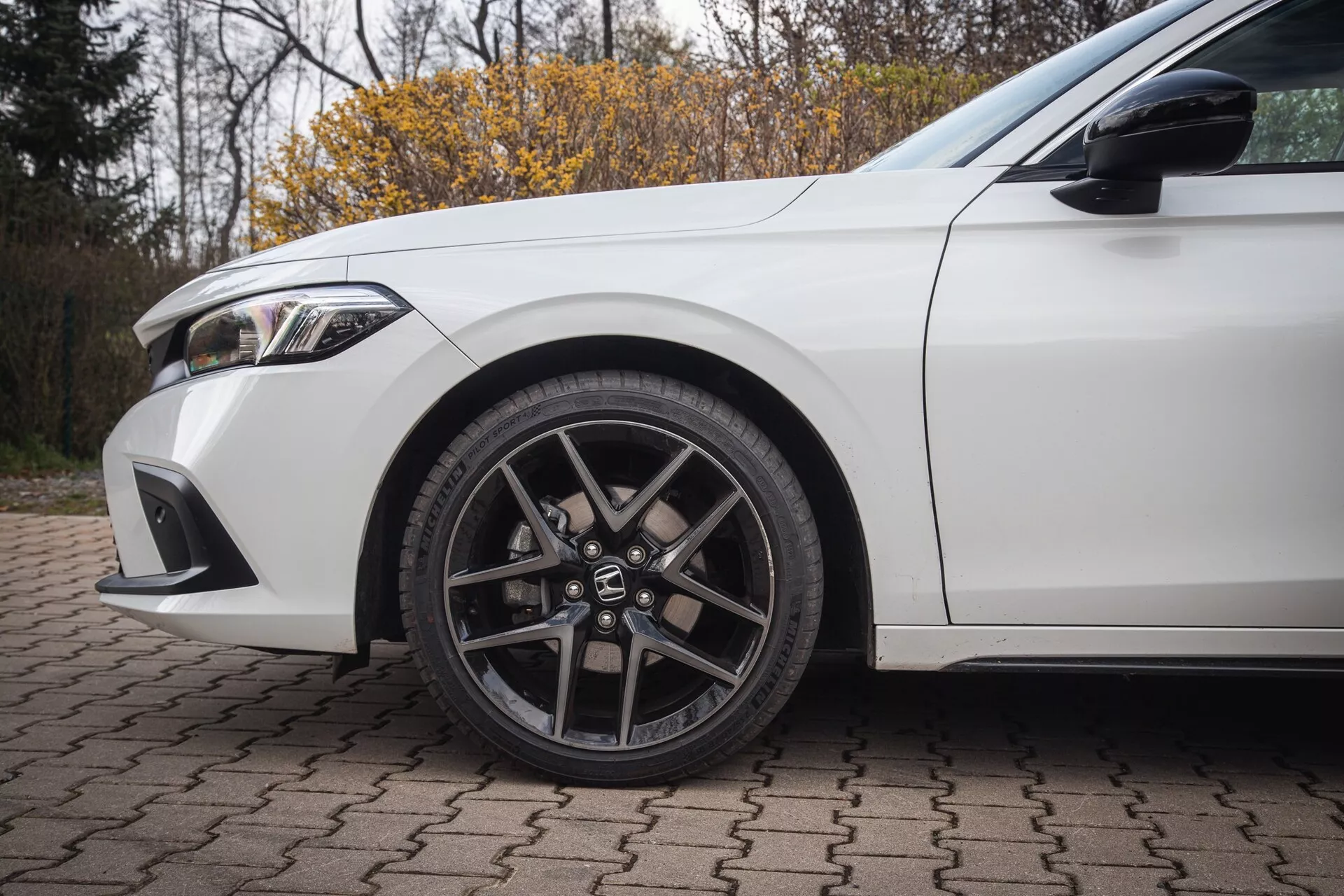 Honda Civic 2022 en blanco, mostrando la rueda delantera izquierda con diseño de aleación de múltiples radios y neumático de perfil bajo, junto con detalles del guardabarros y parte del parachoques delantero en una superficie de ladrillos