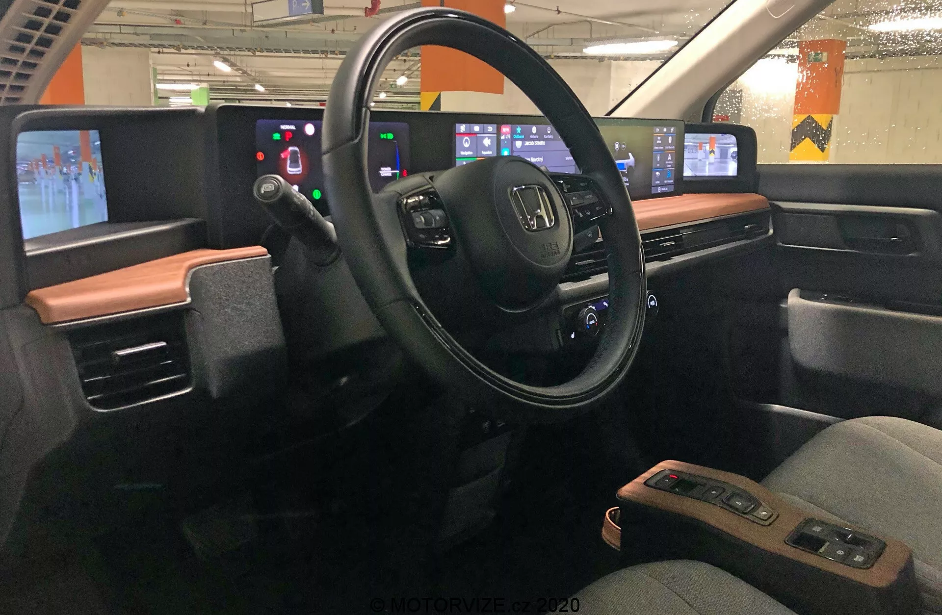 Vista interior del Honda e 2020, enfocándose en el área del conductor vista desde el centro del vehículo. El tablero presenta un diseño moderno con un esquema de color de dos tonos. El volante, con el logo de Honda y varios botones de control, está en el medio, rodeado por un panel de pantalla digital en lugar de indicadores analógicos. A la derecha, la consola central digital incluye paneles táctiles para audio, navegación, control de clima y otras configuraciones. El diseño enfatiza la integración tecnológica, minimizando los botones físicos a favor de interfaces táctiles. El coche parece estar estacionado en un garaje, con luces fluorescentes y pilares de hormigón visibles a través de las ventanas.