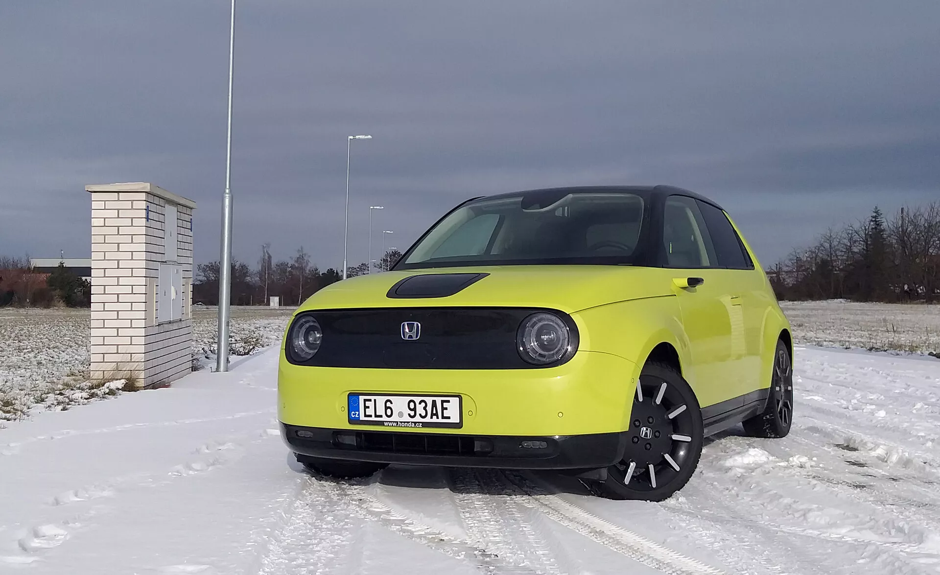 Honda e 2020 de color amarillo brillante sobre una superficie cubierta de nieve con cielo nublado, que presenta faros LED, tecnología de vehículo eléctrico con asistencia avanzada al conductor, llantas de aleación negra y neumáticos de invierno.