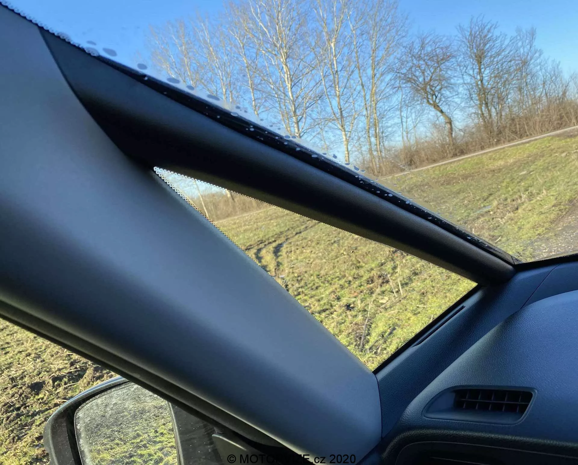 Vista interior de un Honda Jazz Crosstar 2020 mirando hacia la ventana del lado del conductor, enfocándose en el pilar A. El pilar A está cubierto con un material texturizado no metálico y oscurece parcialmente la vista. La ventana lateral muestra gotas de agua, lo que indica lluvia reciente o condensación, con un área de césped y árboles visibles afuera bajo un cielo azul despejado.