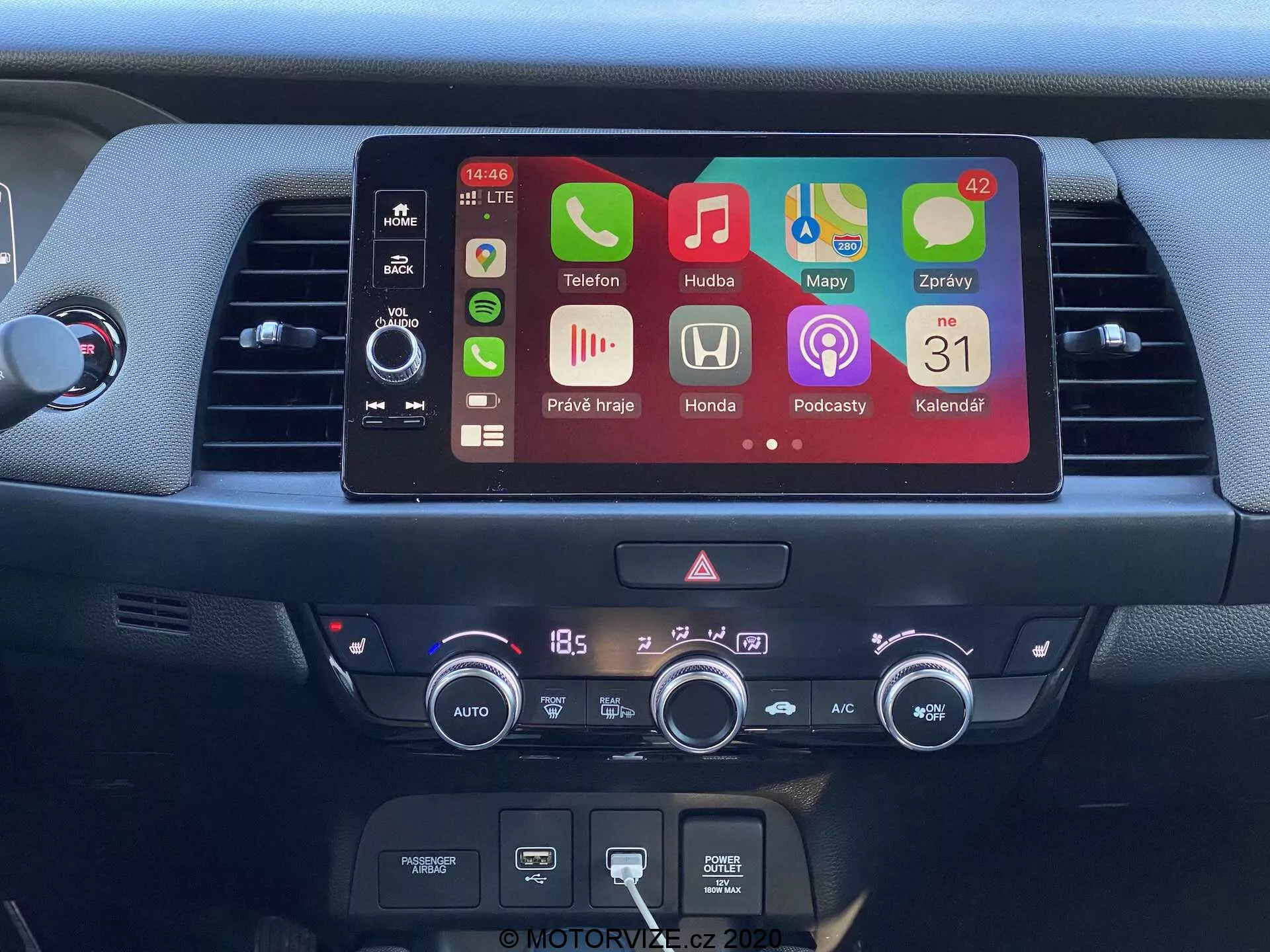 Vista detallada de la consola central del Honda Jazz 2020, que muestra una moderna pantalla de infoentretenimiento táctil con iconos de aplicaciones coloridas incluyendo teléfono, música, navegación, configuraciones del coche, podcasts y calendario. Debajo de la pantalla se encuentran controles físicos del sistema de audio, una ranura para CDs y un sistema de control climático digital ajustado a 18.5 grados Celsius. La imagen resalta características avanzadas como conectividad Bluetooth, integración de smartphones y control automático del clima.