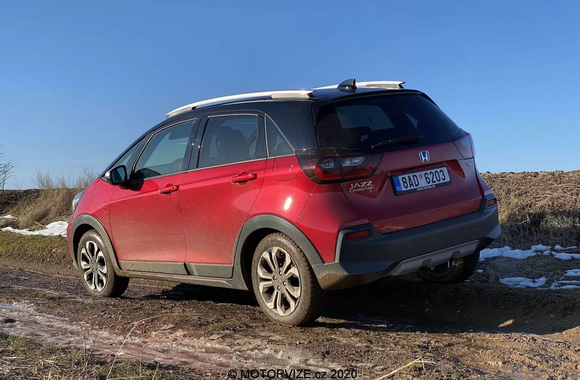 Un SUV Honda Jazz Crosstar rojo de 2020 visto desde una perspectiva trasera de tres cuartos, mostrando detalles como insignias de marca y modelo, luces traseras LED, parachoques trasero con arañazos y sensores, llantas de aleación, neumáticos para todo tipo de clima, rieles de techo, antena de aleta de tiburón, ventanas tintadas con líneas de descongelación y una matrícula centrada. La apertura del maletero tipo hatchback y parches de nieve en el suelo con un campo seco en el fondo sugieren un entorno de últimos otoño, invierno o principios de primavera bajo un cielo despejado al mediodía o primeras horas de la tarde.