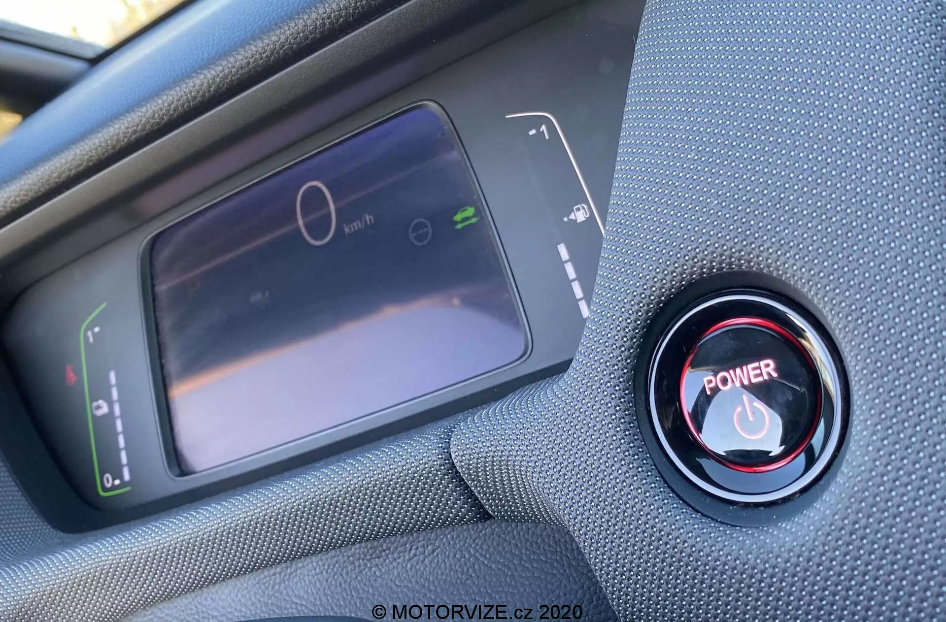 Vista interior de cerca desde la perspectiva del conductor de un Honda Jazz Crosstar 2020, centrada en la pantalla del cuadro de instrumentos digital y el botón de arranque/parada del motor rojo. La pantalla LCD a la izquierda muestra un velocímetro estacionario y un gráfico de uso de energía, mientras que a la derecha, el botón de arranque/parada rojo está rodeado por un anillo metálico. La foto está tomada en un ángulo oblicuo desde arriba, con un enfoque nítido en el botón y un enfoque suave en la pantalla.