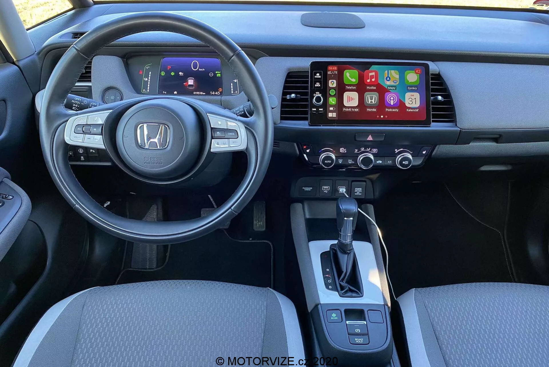 Vista interior del Honda Jazz Crosstar 2020 desde la perspectiva del conductor. Se destaca un volante multifuncional moderno con el logo de Honda, que incluye controles para audio, control de crucero y comandos de voz. Detrás del volante hay un panel de instrumentos digital que muestra información esencial. La consola central incluye un sistema de infoentretenimiento que probablemente soporte Apple CarPlay o Android Auto, con controles de clima debajo. Palanca de cambios simple y controles adicionales para el freno de mano electrónico y la selección de modos de conducción. La iluminación y el ángulo destacan la comodidad y el diseño ergonómico.
