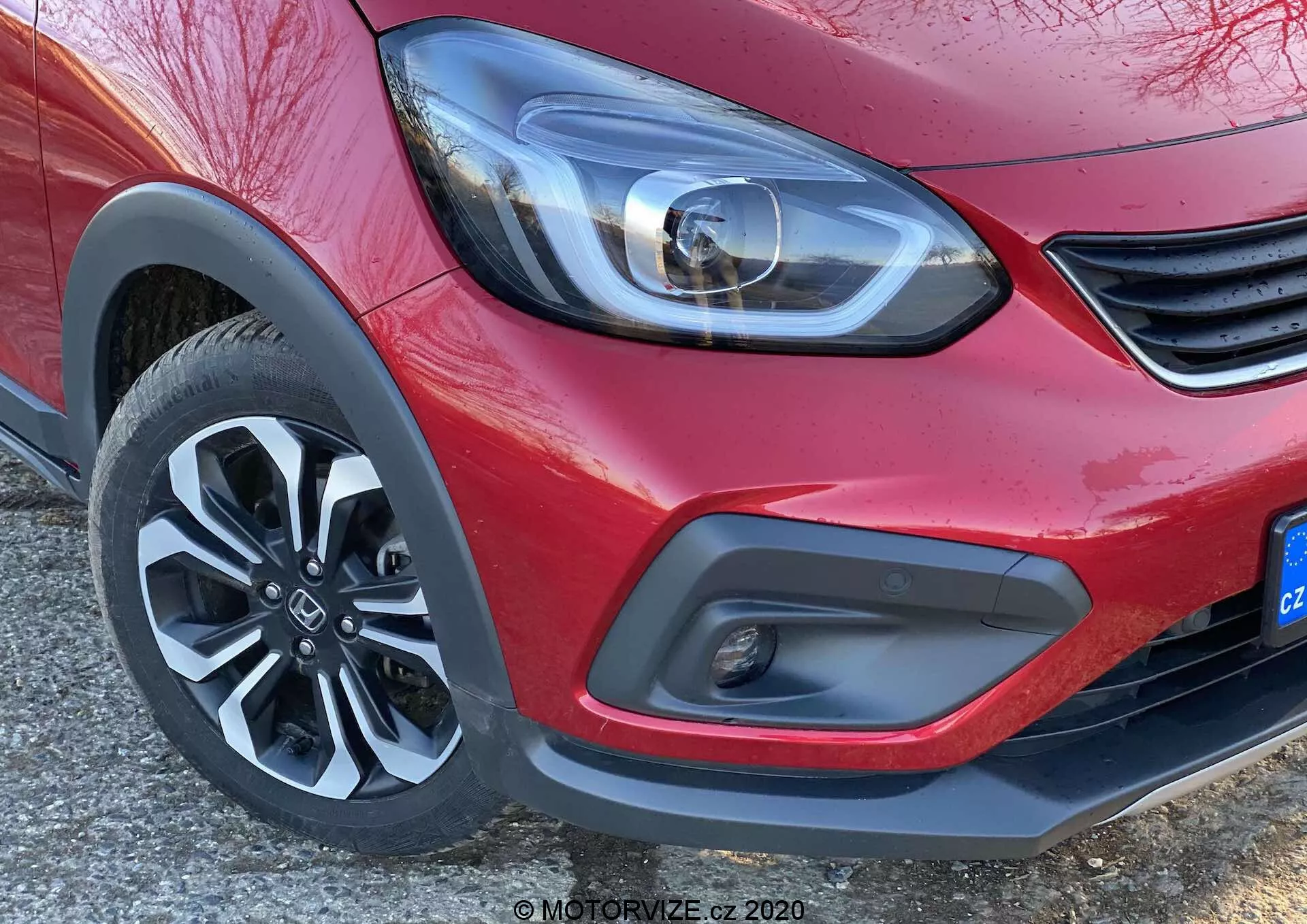 Vista en primer plano de la esquina delantera de un Honda Jazz 2020 (Jazz Crosstar) rojo, enfocándose en el moderno faro LED, el detalle minimalista de la rejilla, el parachoques delantero bicolor con luz antiniebla y la rueda de aleación de cinco radios dinámica.